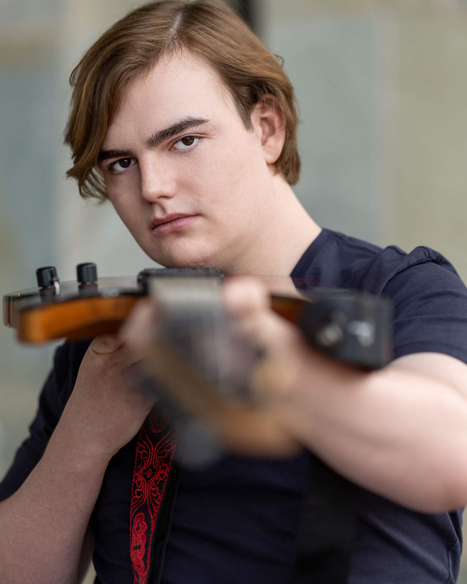Young male musician with brown hair holding guitar toward camera wearing dark t-shirt.