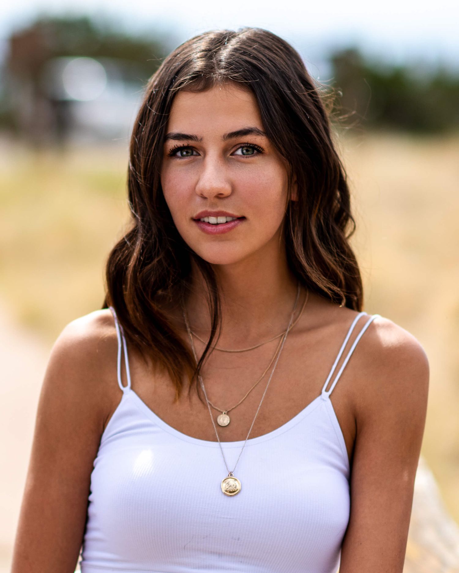 Brunette woman in white camisole with layered gold necklaces posing outdoors in a sunny field portrait.