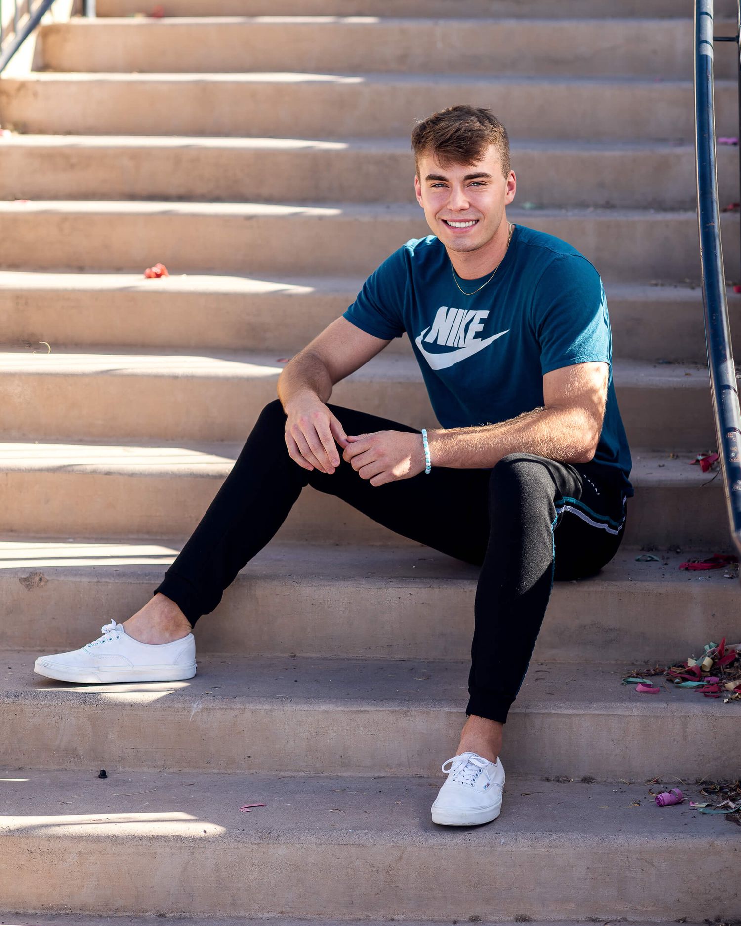 Young man in Nike t-shirt and black jeans sitting on outdoor steps, smiling, wearing white sneakers.