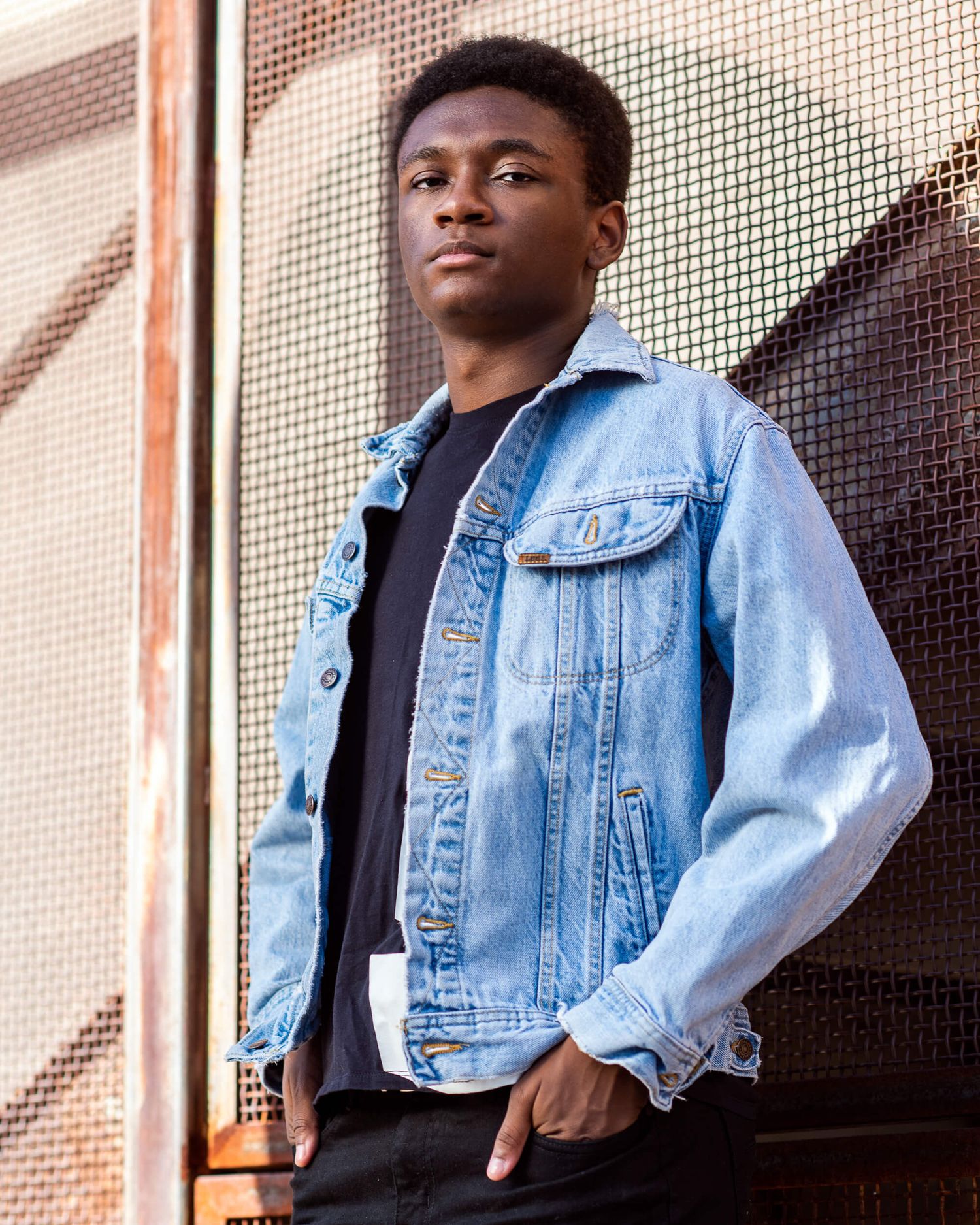Young man in light wash denim jacket and black shirt posing against metal mesh wall outdoors.
