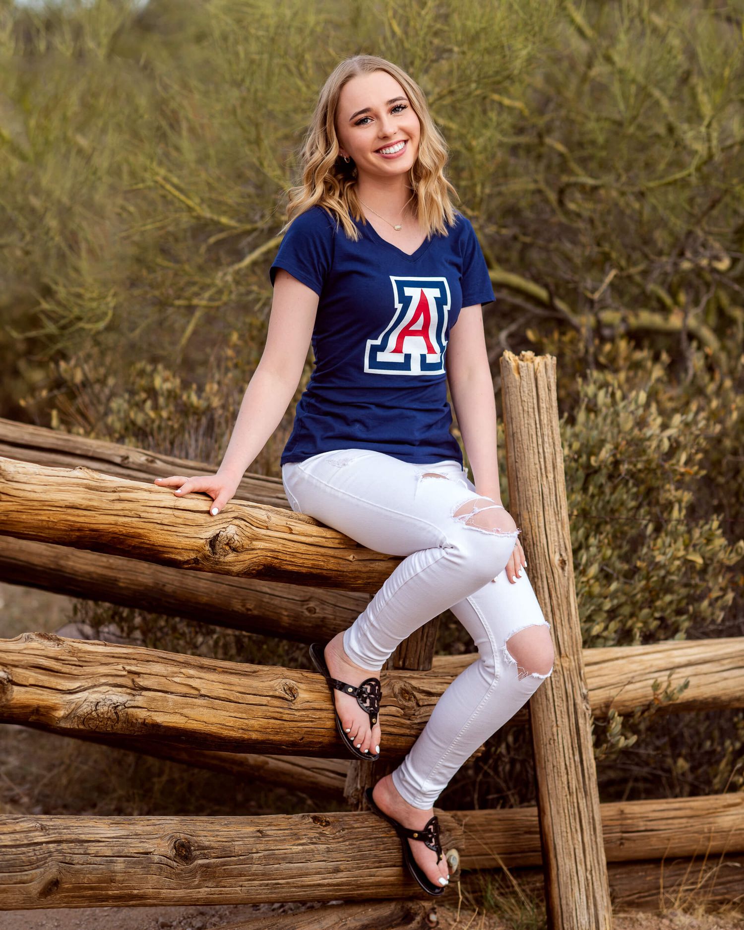 Smiling blonde teen girl senior photo wearing University of Arizona shirt on wooden fence.