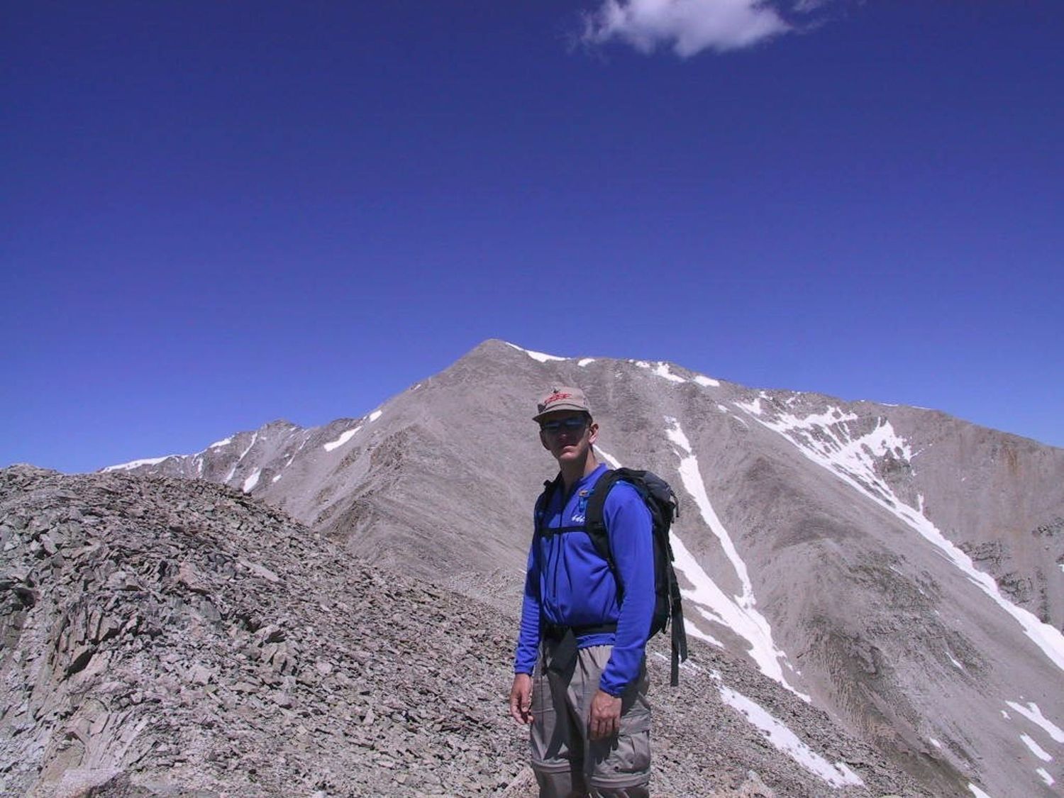 Hiker in blue jacket standing on rocky mountain ridge under clear blue sky.