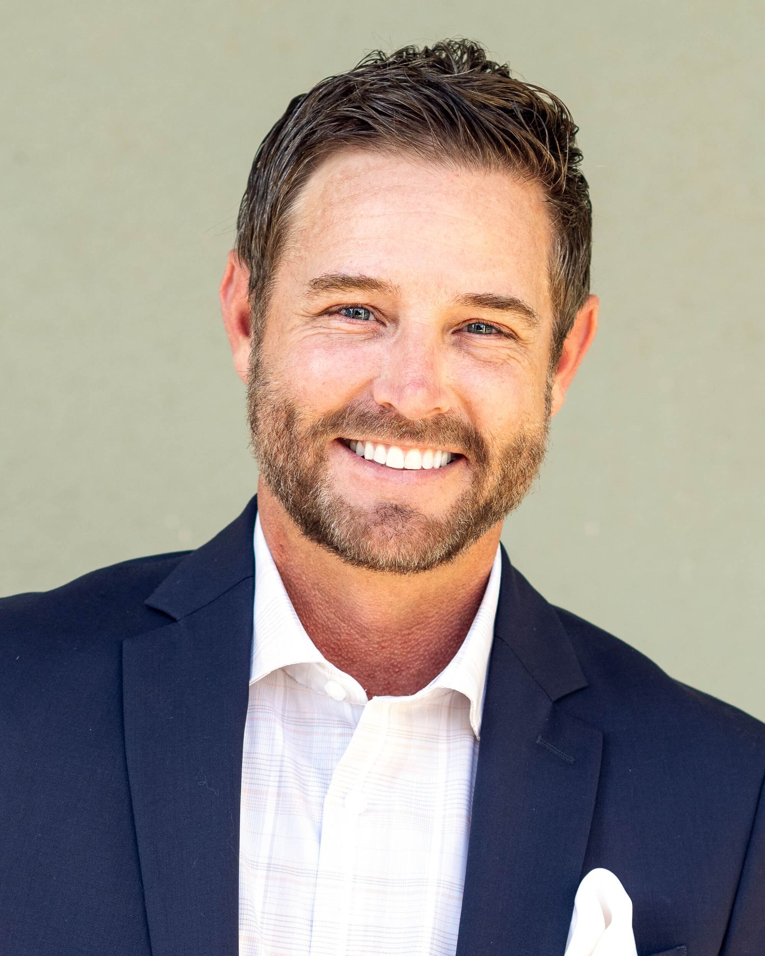 Smiling man with brown beard wearing a navy blazer and white shirt against a green background.