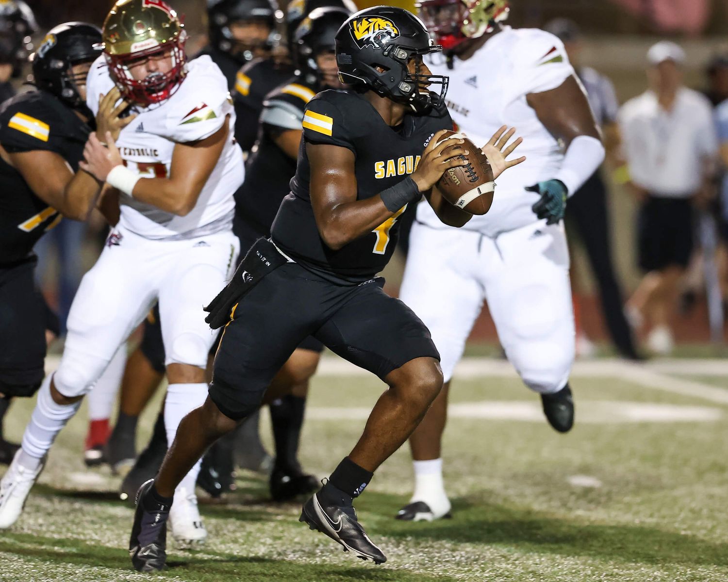 Football quarterback #1 in black uniform scrambles during a night game under lights.
