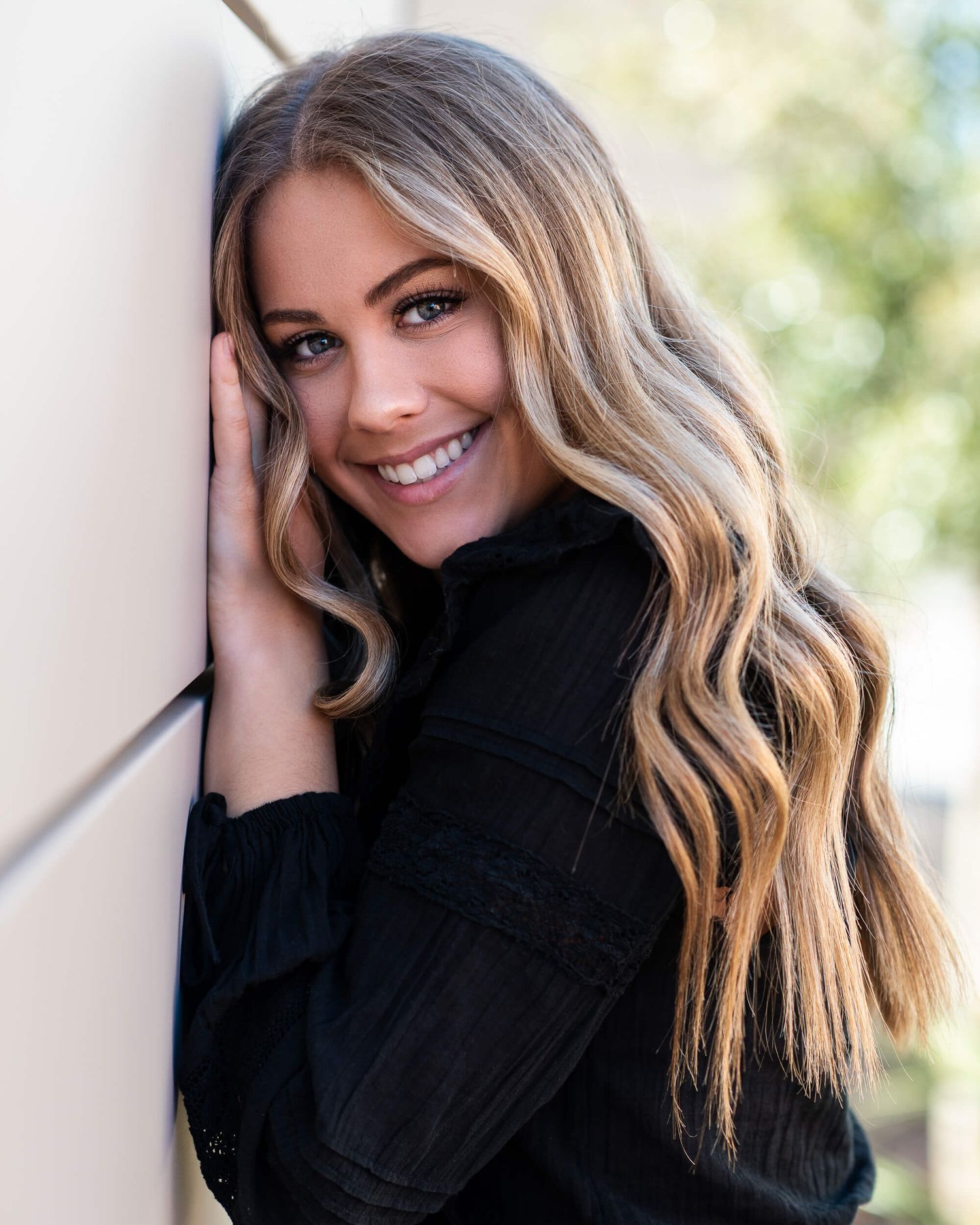 Blonde woman with wavy hair smiles warmly leaning against a wall in a black long-sleeve top outdoors.