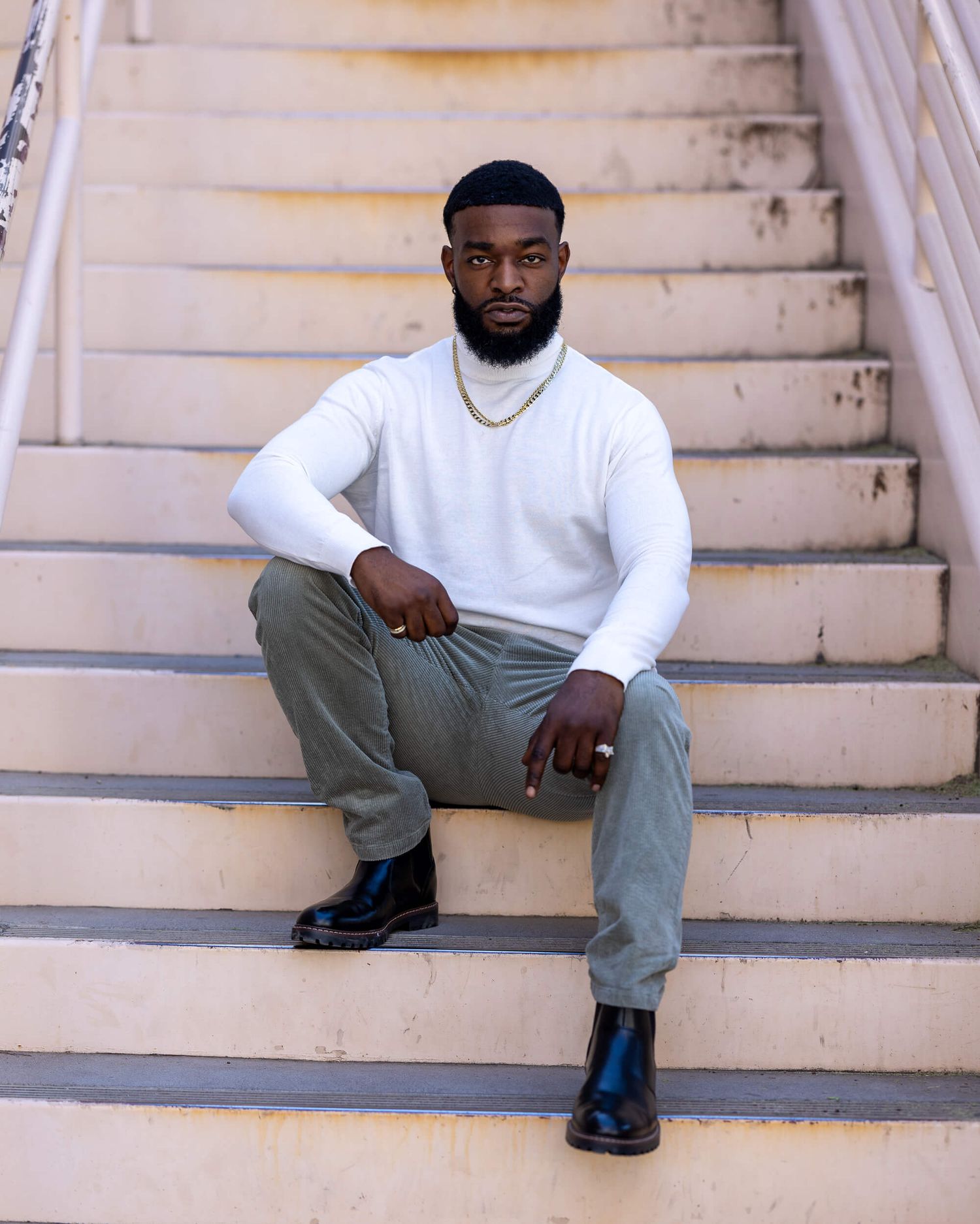 Man in white sweater and olive pants sitting on beige steps wearing black boots.