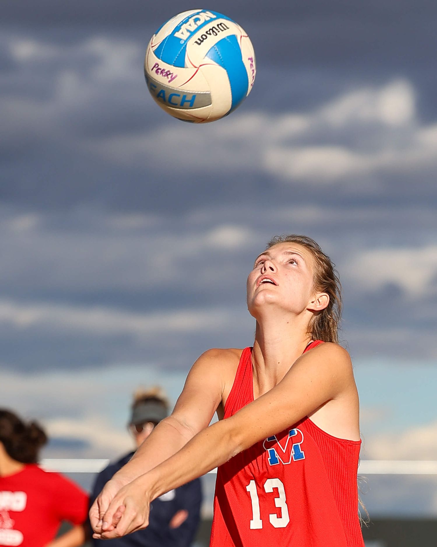 Female beach volleyball player in red number 13 jersey bumps the ball while looking up during outdoor play.