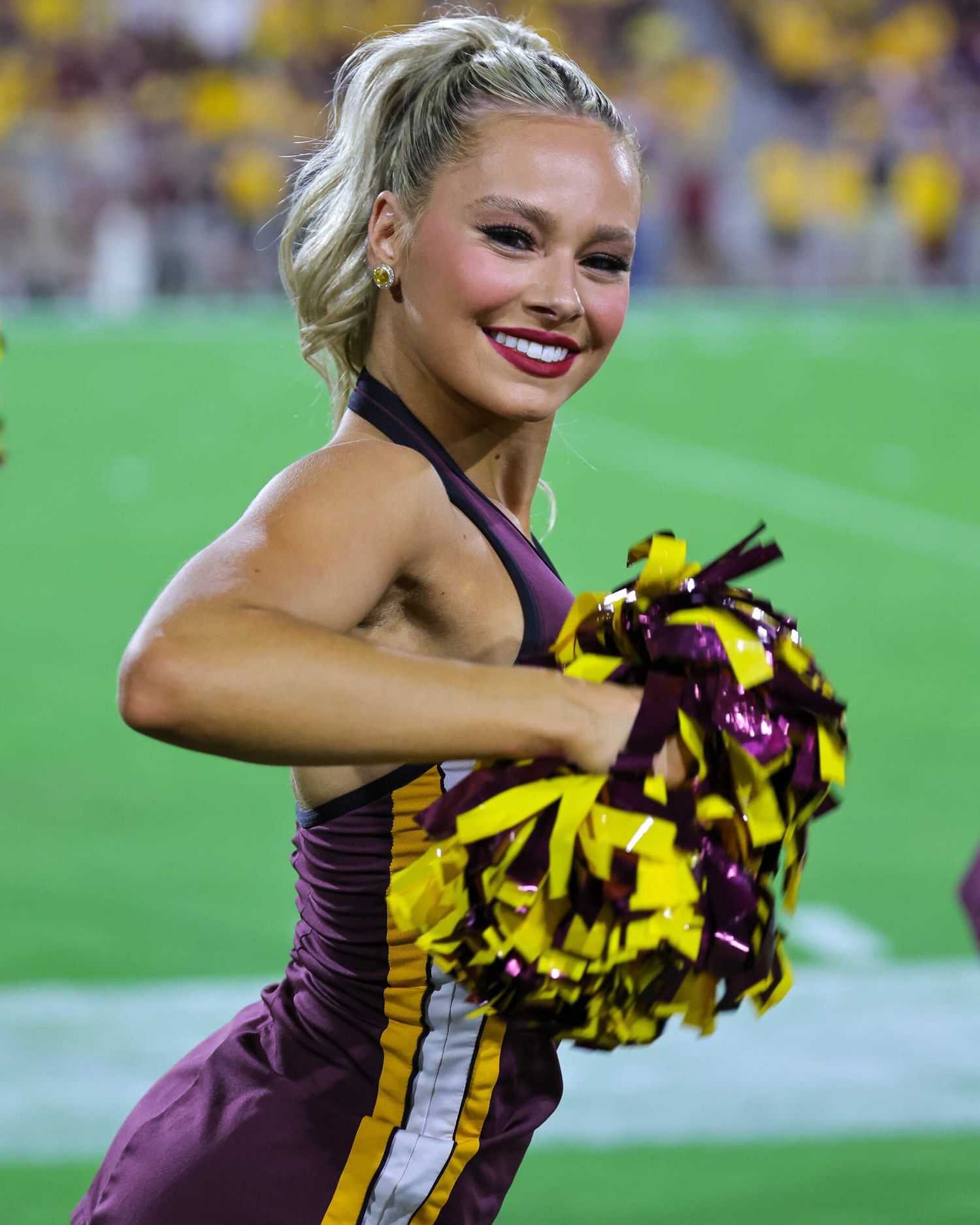 ASU cheerleader in maroon uniform smiles while holding gold and maroon pom-poms at a college football game.