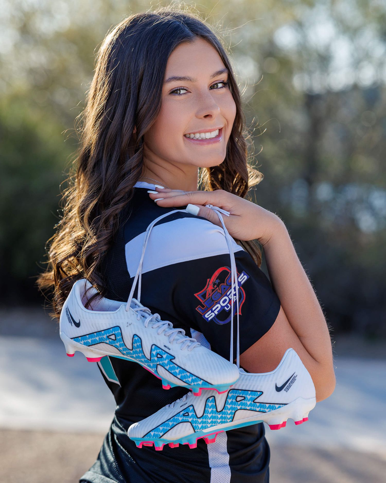 Brunette teen girl senior photo holding Nike cleats wearing sports jersey outdoors.