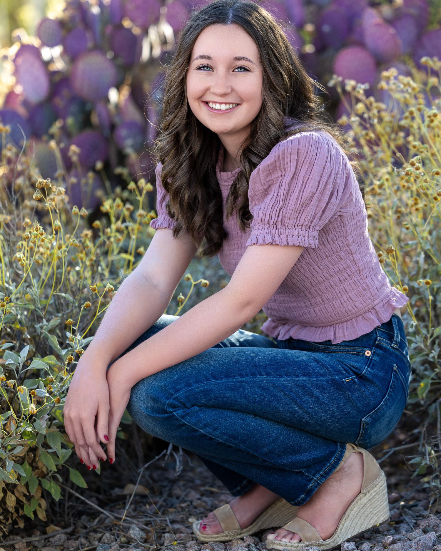 Young woman in pink top and jeans crouching among purple wildflowers, smiling outdoors in a garden setting.