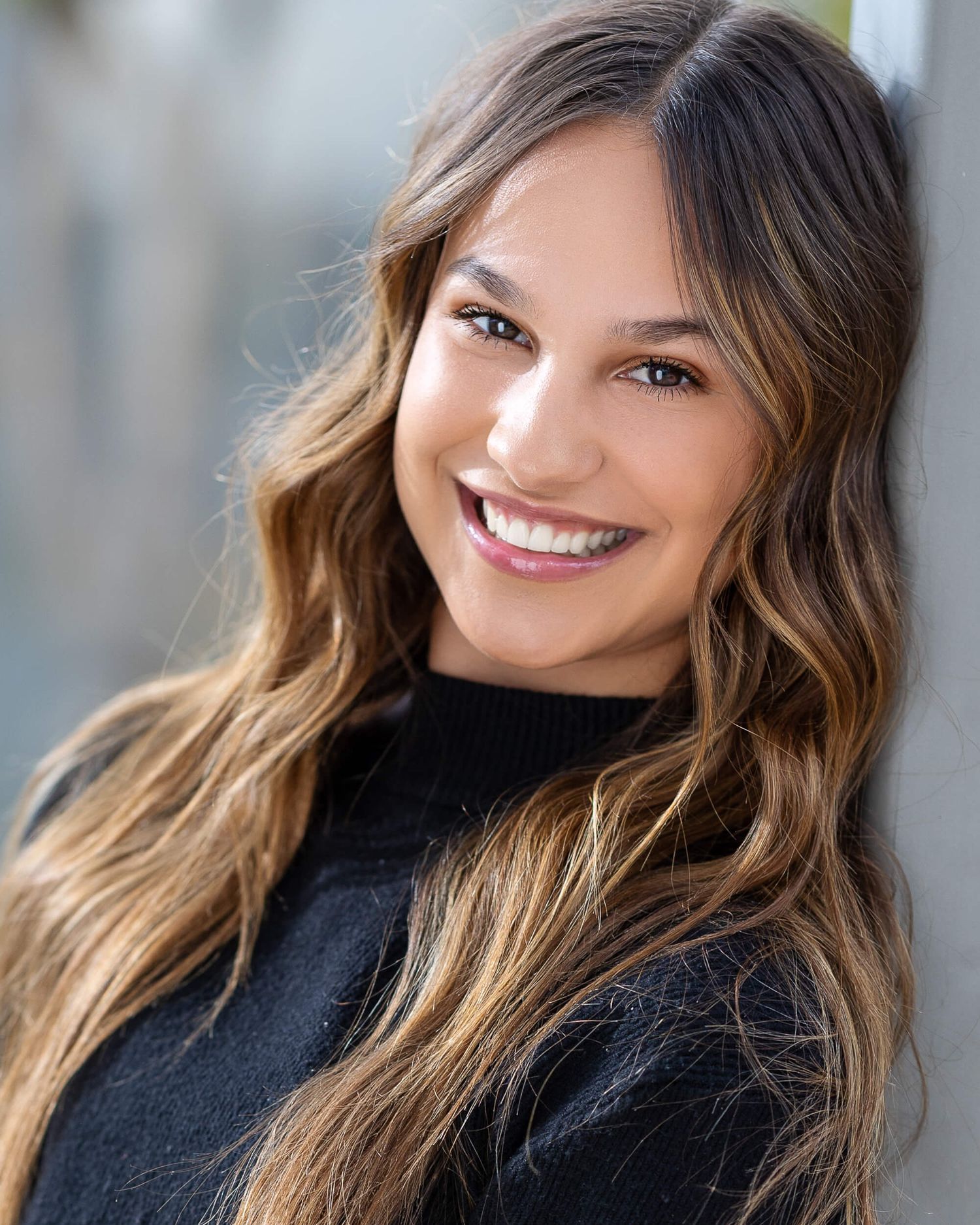 Brunette woman with highlighted wavy hair smiles brightly in black turtleneck headshot.