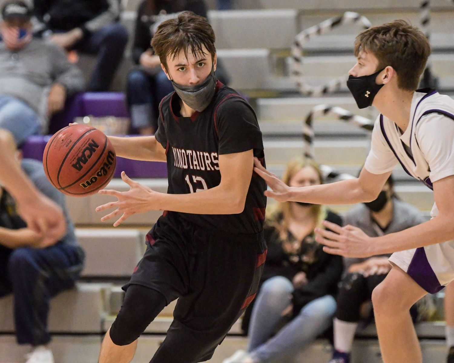 High school basketball player in black Minotaurs jersey dribbles past a masked defender.