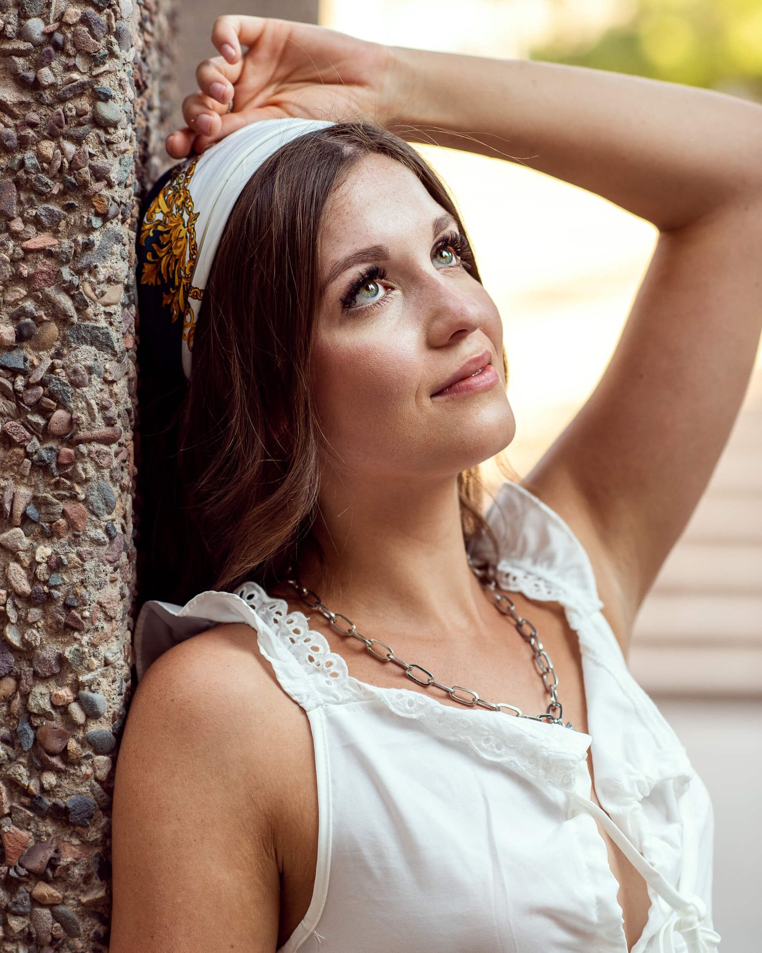 Dark-haired woman in white sleeveless top leans against stone pillar looking upward outdoors.