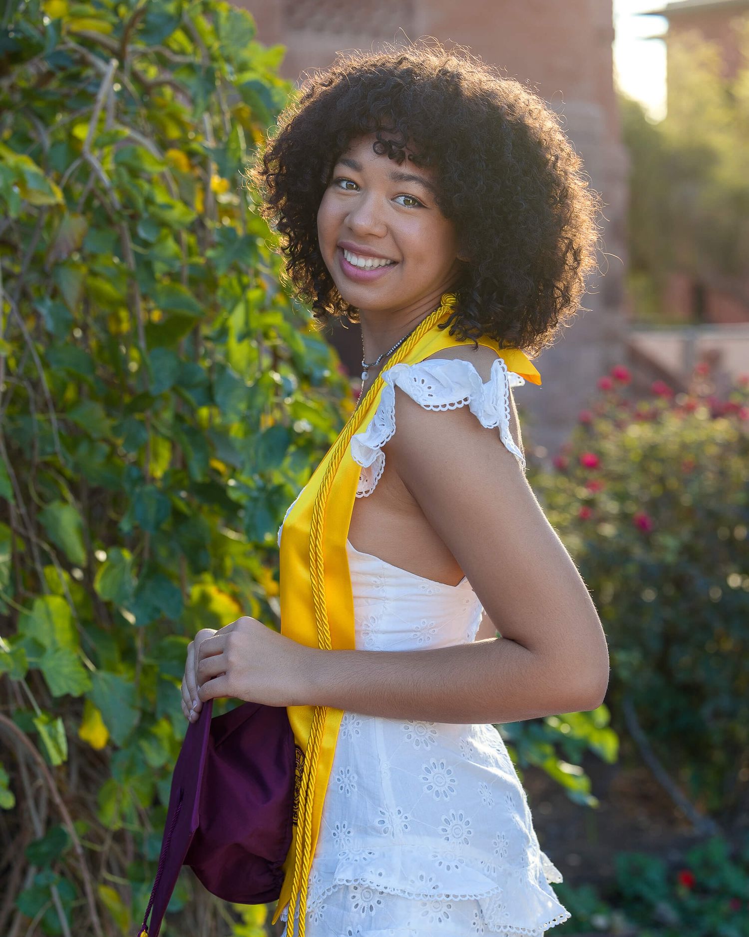 Smiling young woman in white ruffled top with gold graduation stole standing beside green garden foliage.