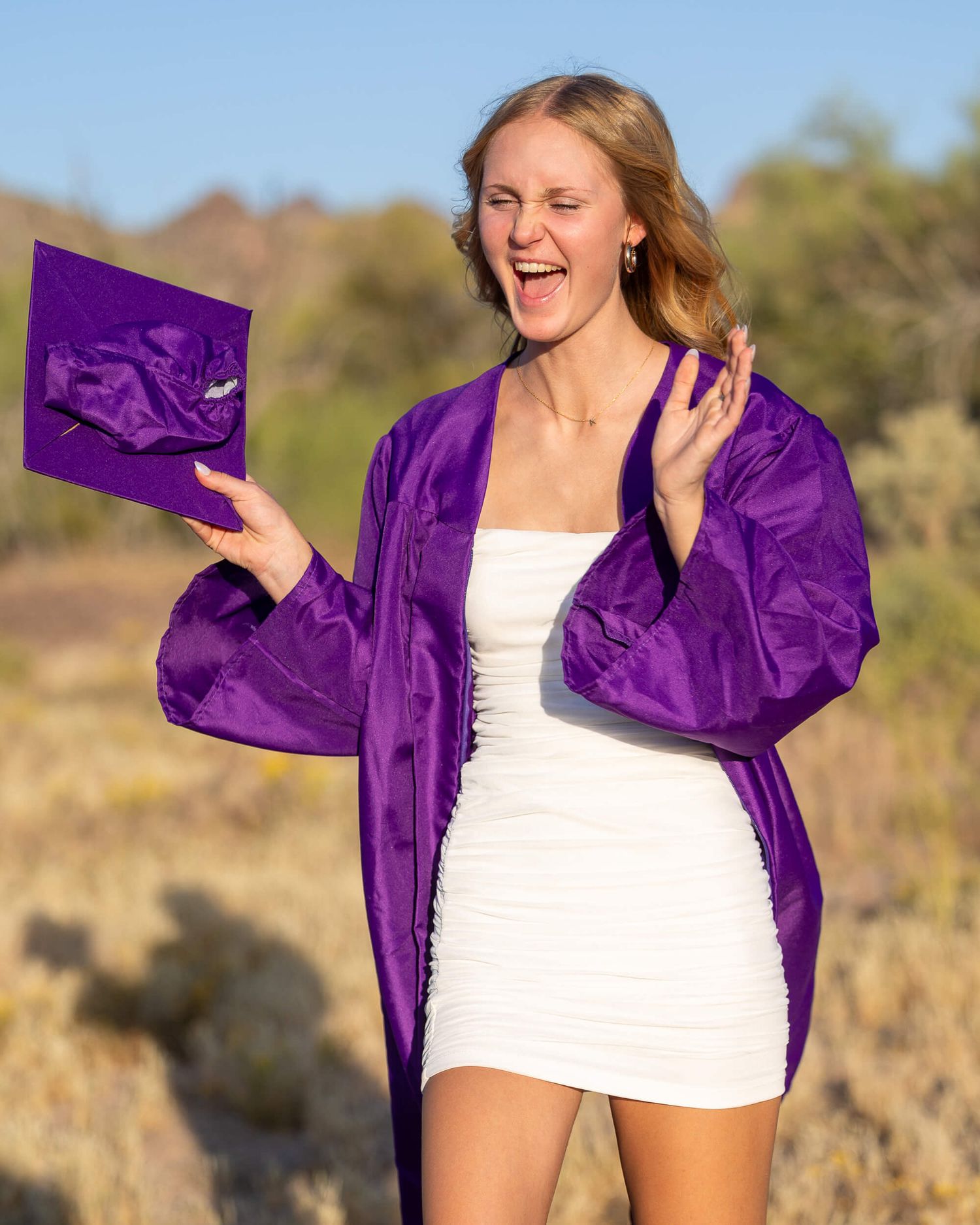 Young woman in purple graduation gown and cap laughing outdoors, wearing white dress, celebrating graduation milestone.