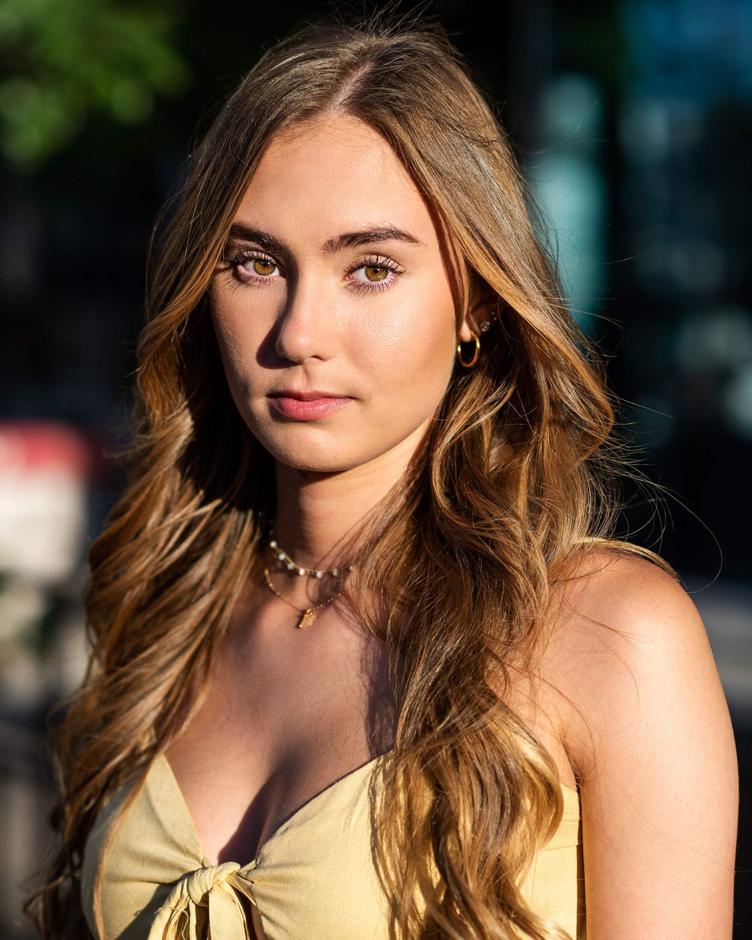 Young woman with wavy brown hair in yellow top wearing gold earrings and pearl necklace outdoors.