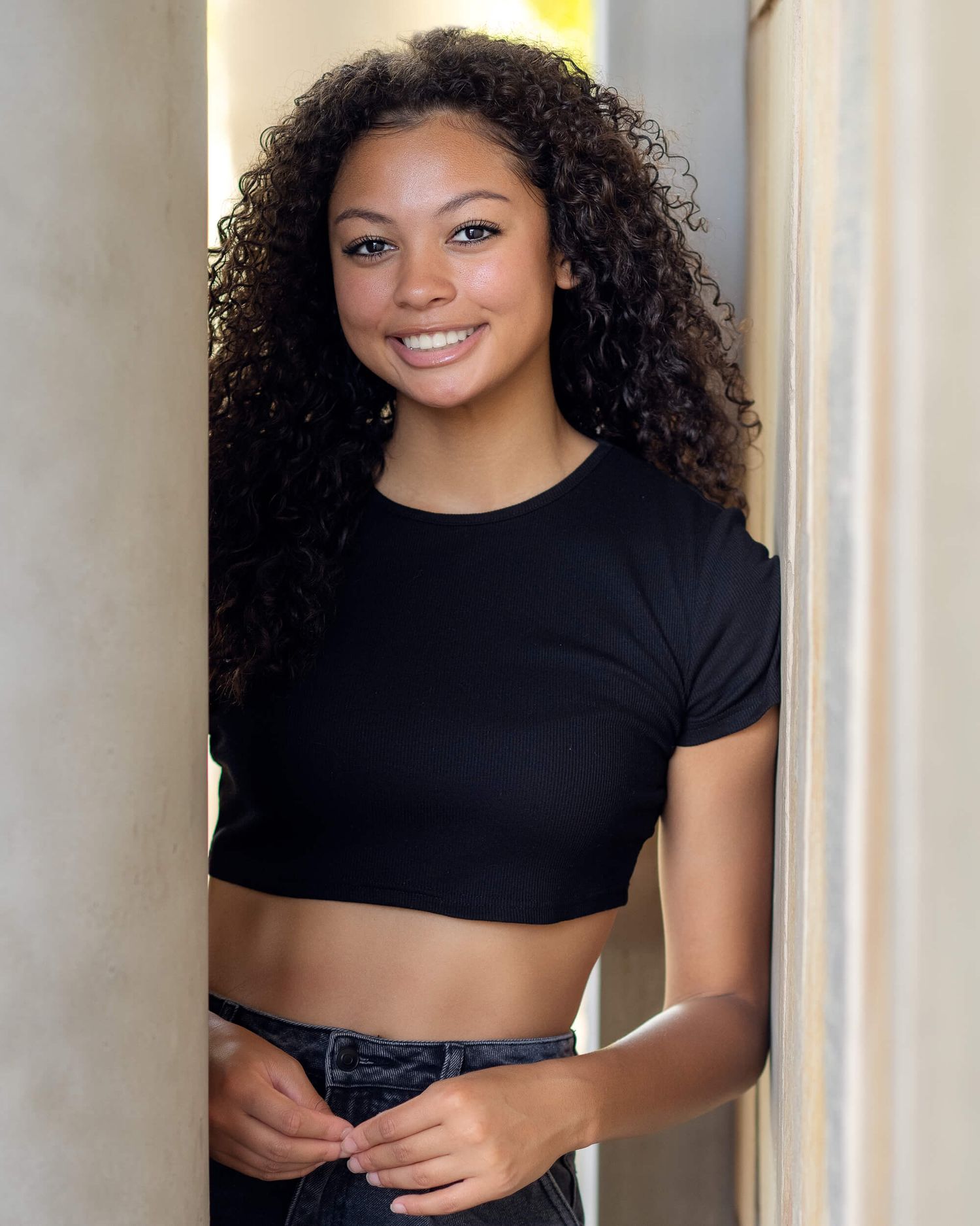 Young woman with curly hair smiling in black crop top leaning against wall.