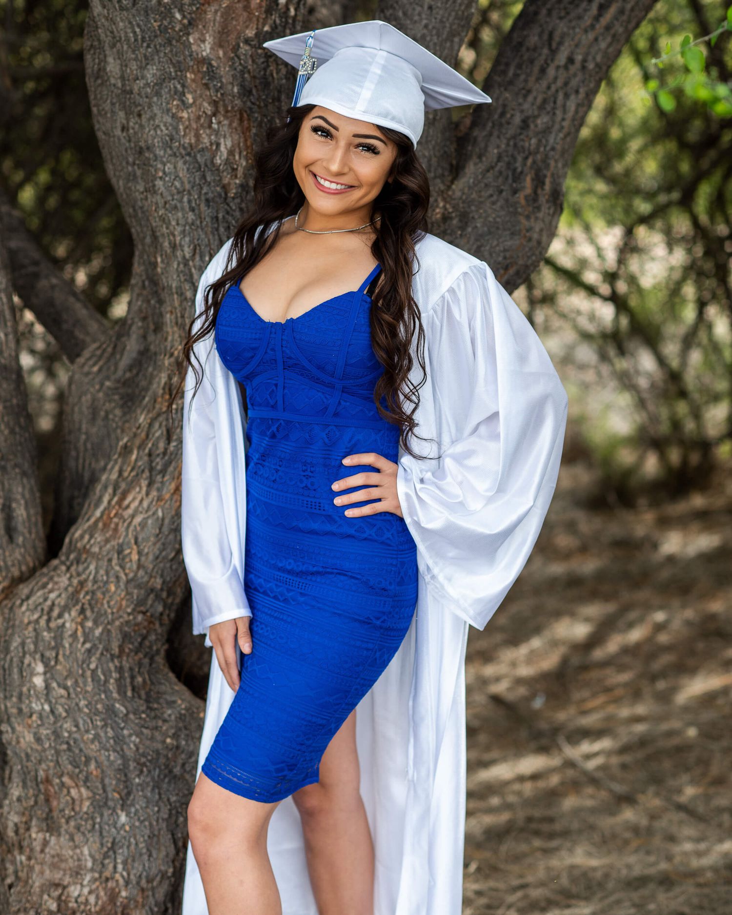 Young woman in white graduation cap and gown over blue dress smiling outdoors among trees.