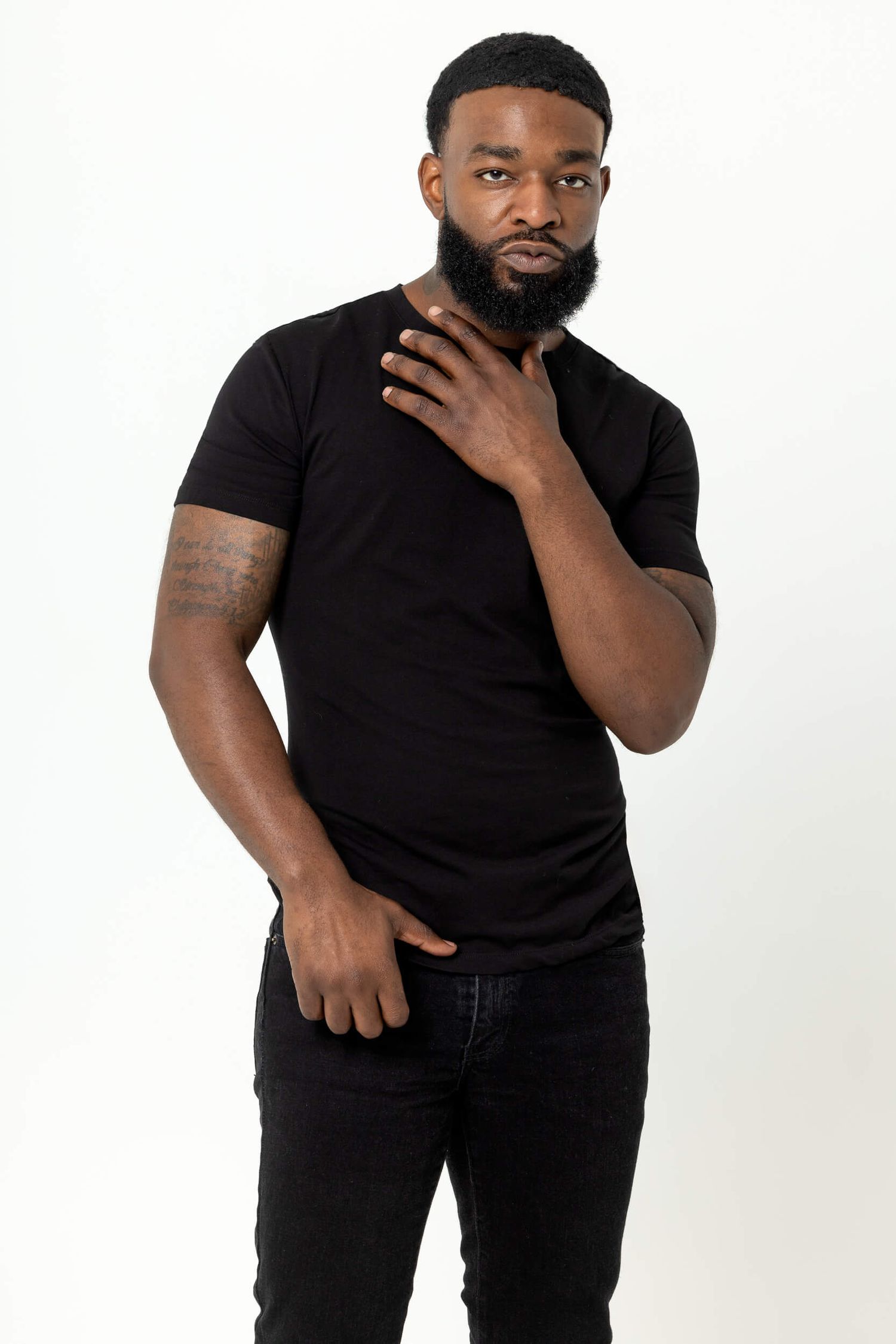 Black man in black t-shirt and jeans posing with hand on chest against white background.