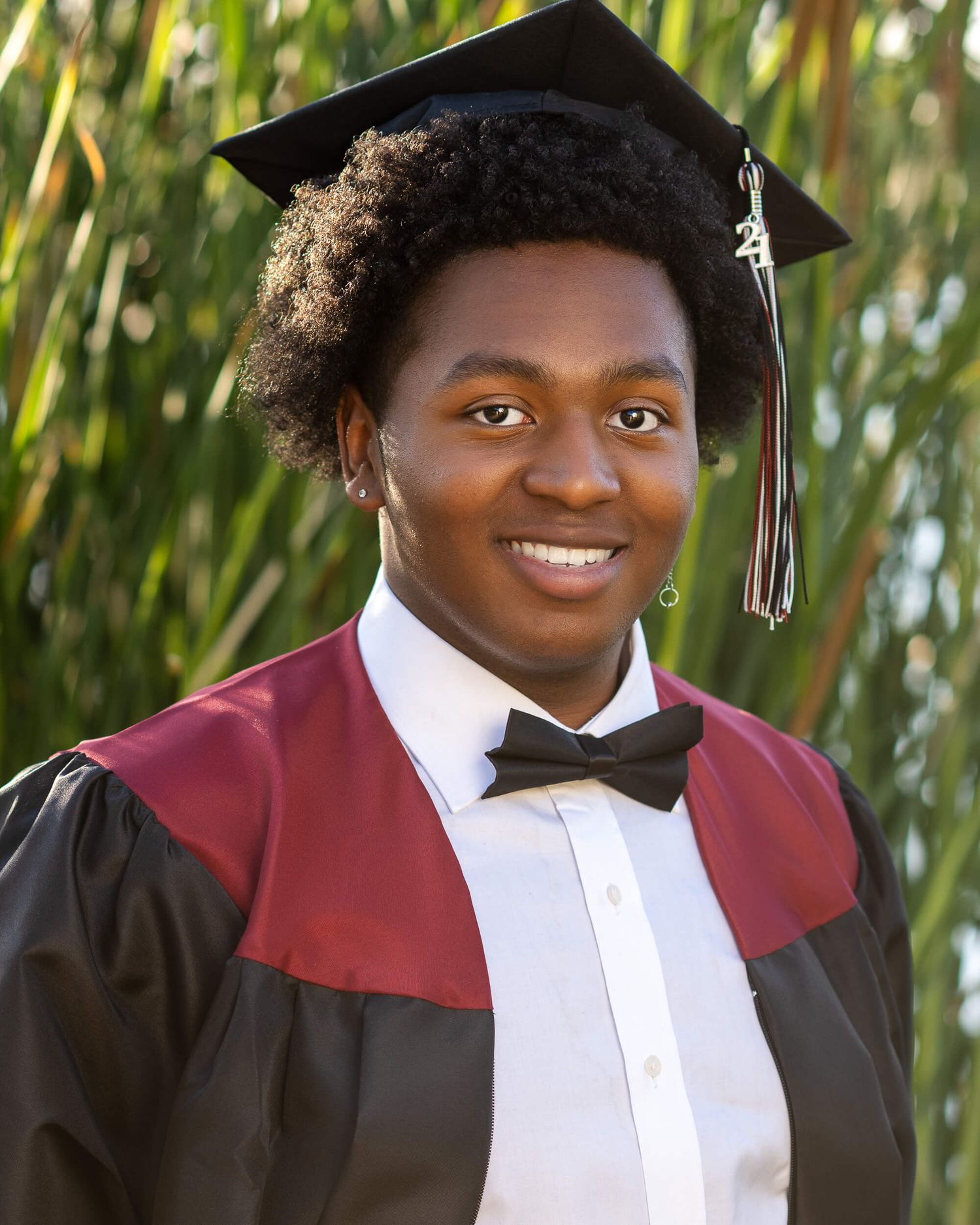 Smiling graduate wearing black cap and gown with red hood and bow tie outdoors.