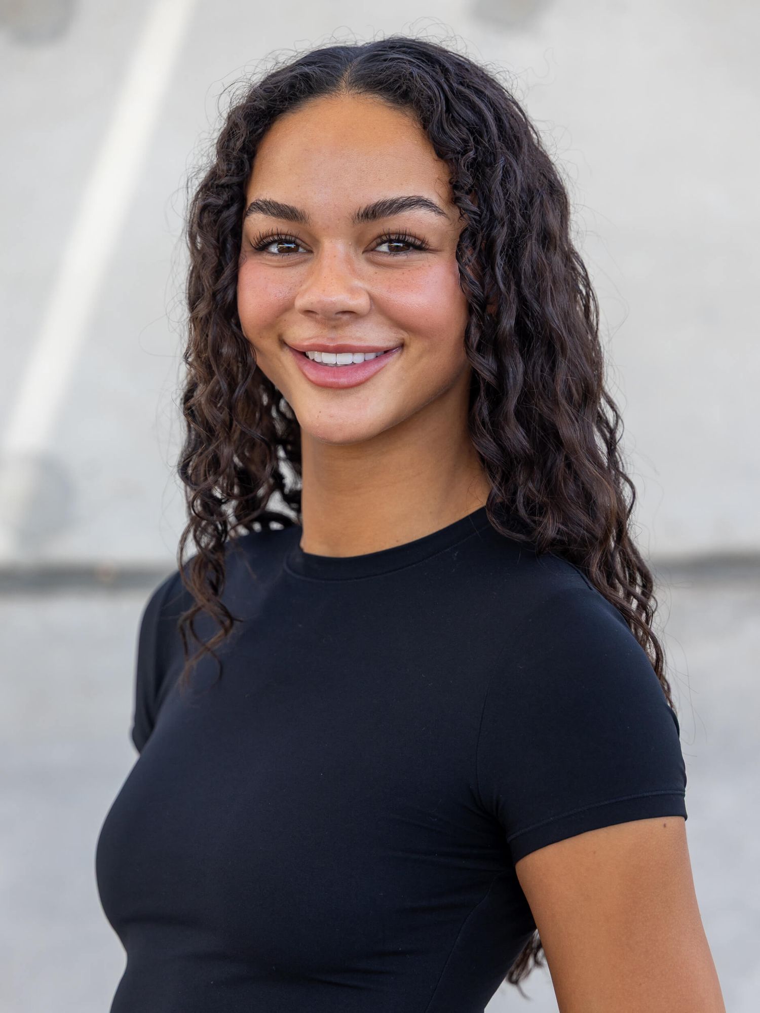 Young woman with curly dark hair smiling confidently, wearing a black fitted t-shirt in an outdoor setting.