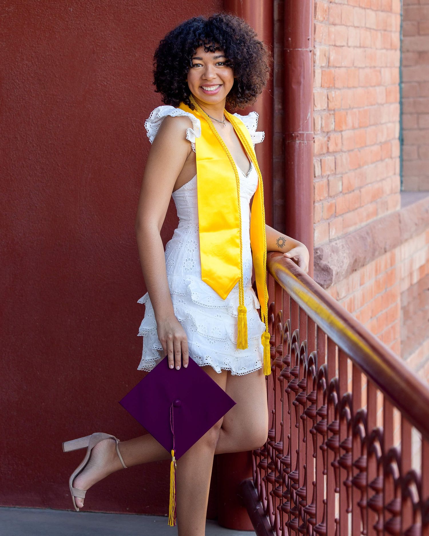 Smiling graduate in white dress with gold stole posing on red brick building staircase railing.
