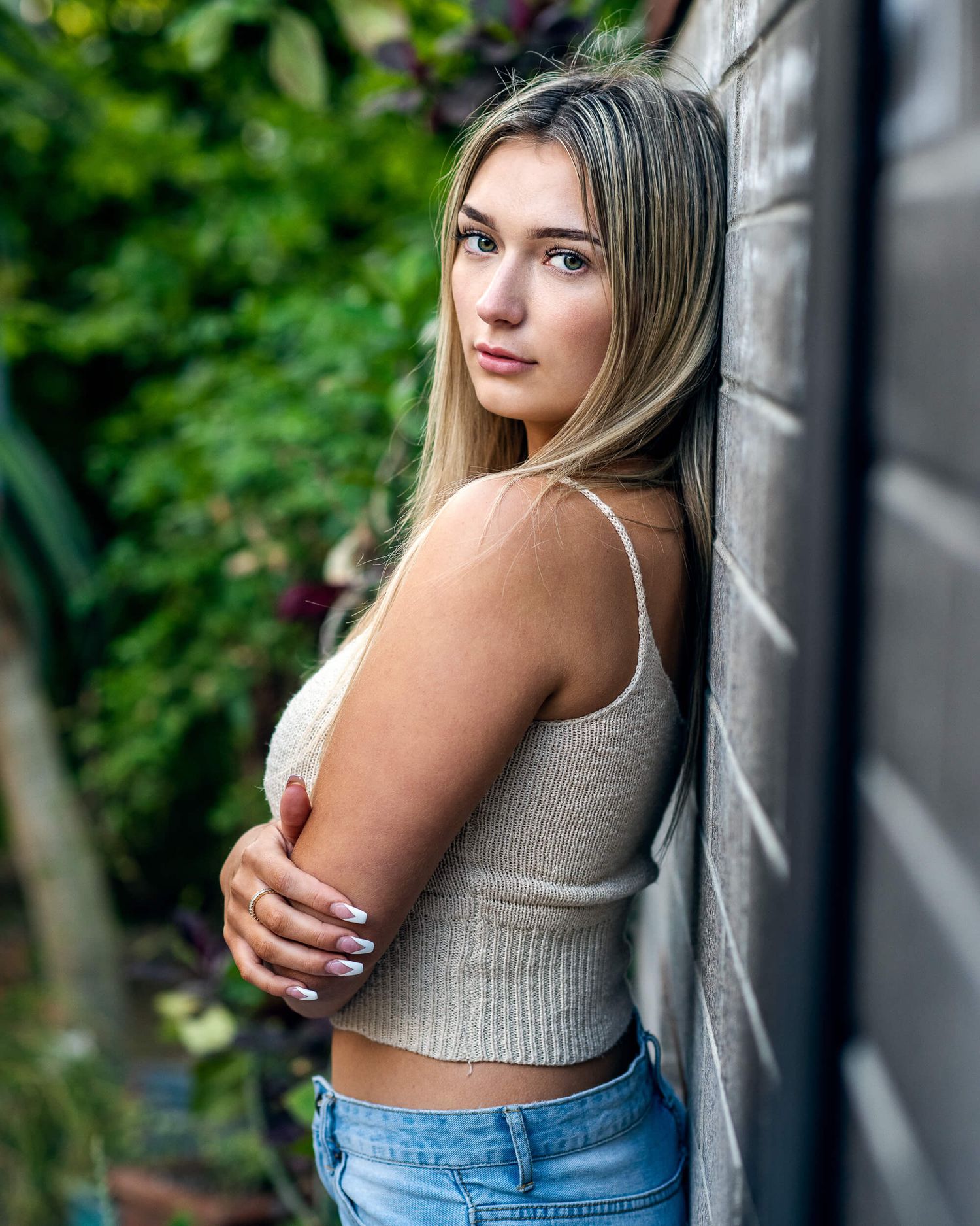 Young blonde woman in beige crop top and jeans leans against brick wall outdoors.