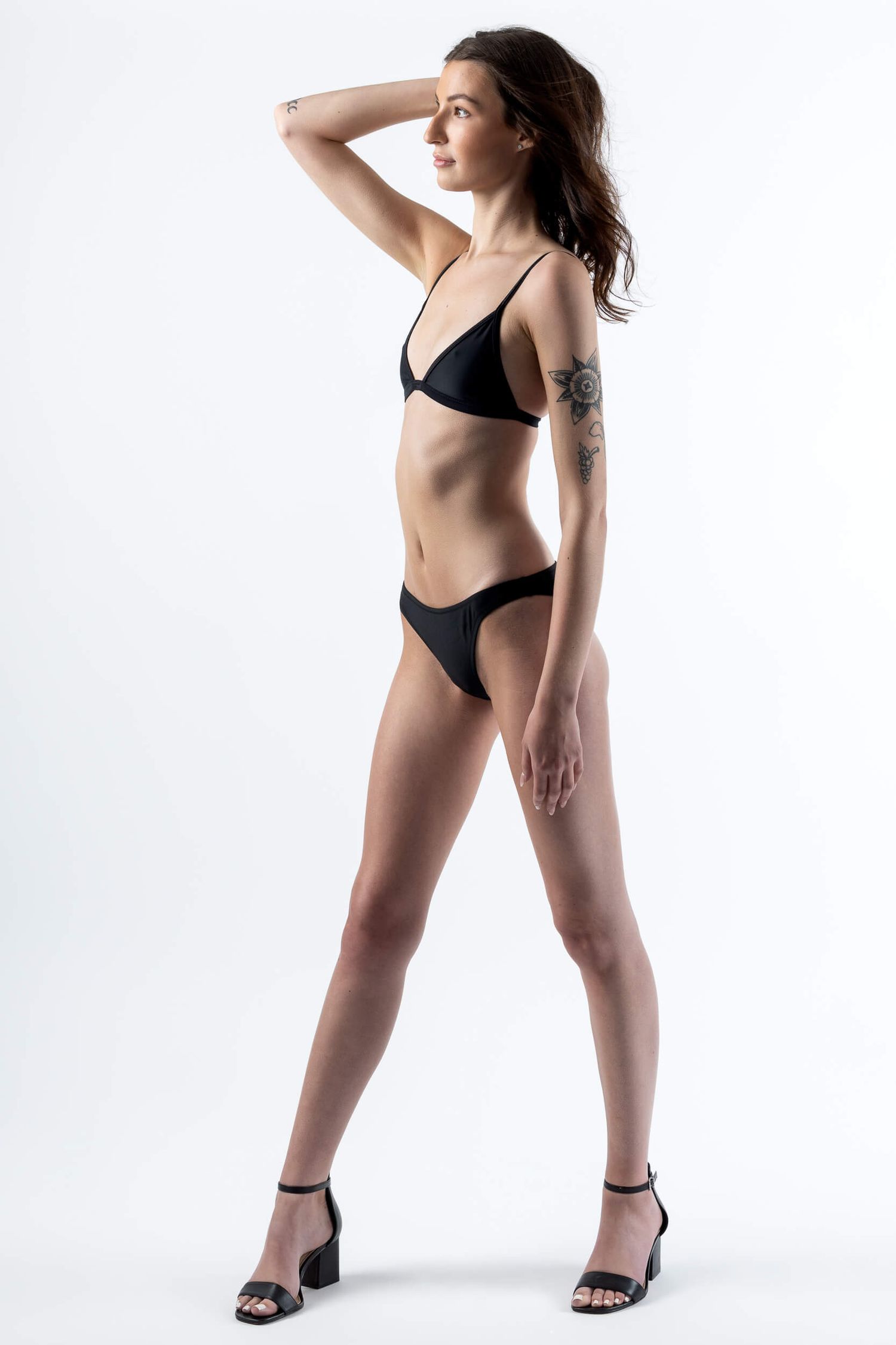 Slender woman in black bikini with arm tattoo posing in heels on white background.