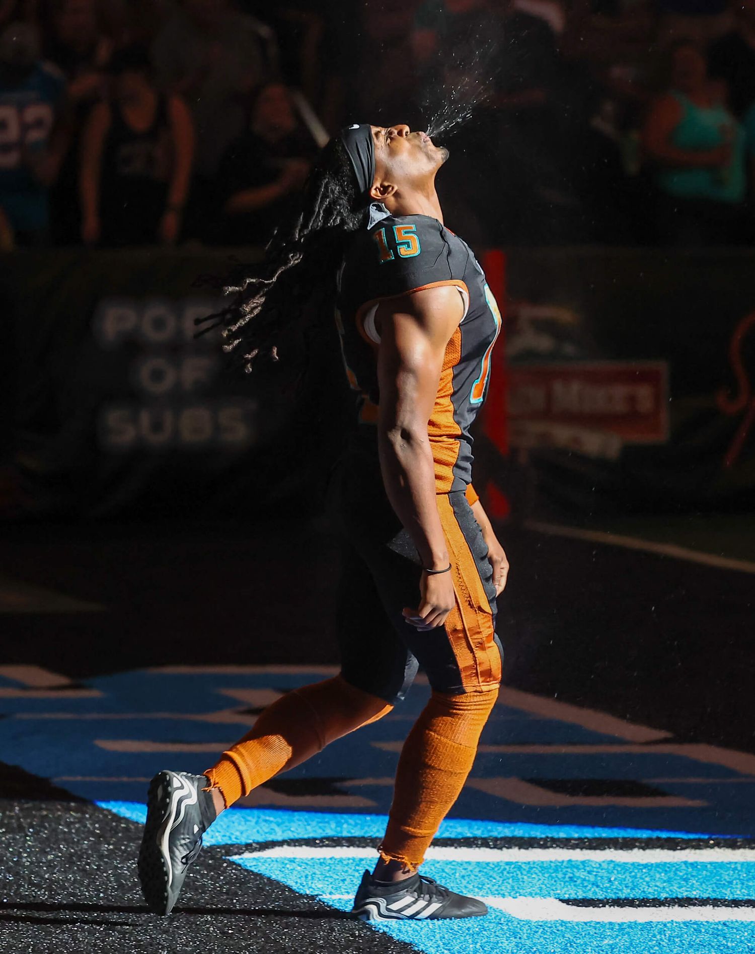 Football player in orange uniform spits water dramatically under bright stadium lights.