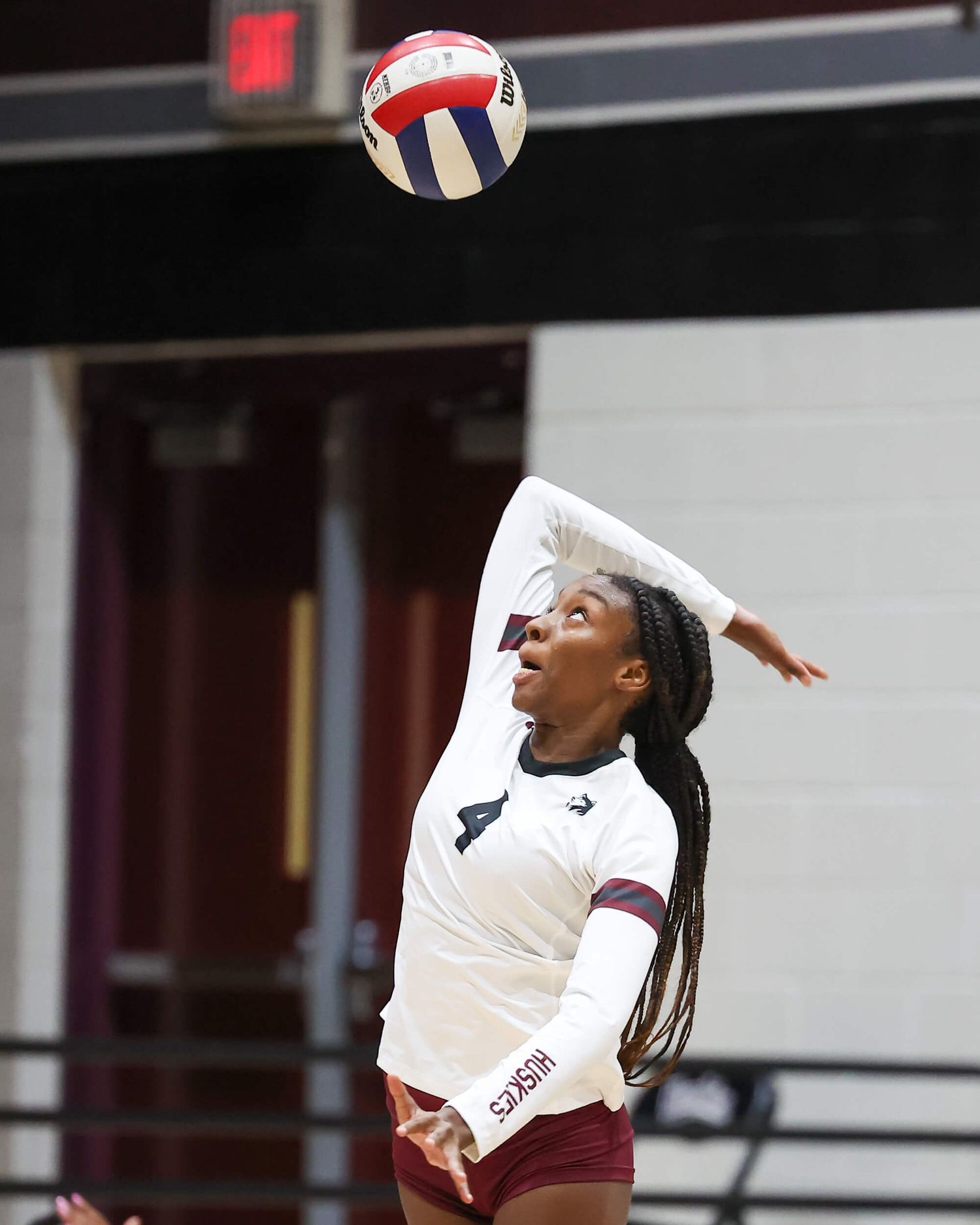 Female volleyball player in maroon jersey number 4 leaps to spike the ball during an indoor match.