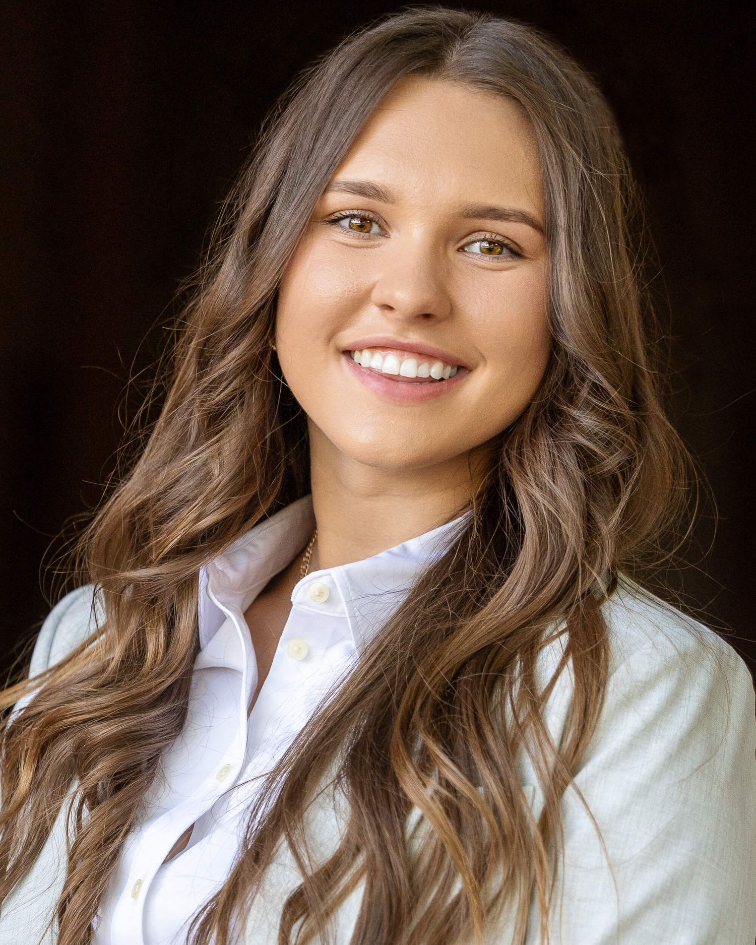 Brunette woman with long wavy hair smiles warmly in white shirt professional headshot.