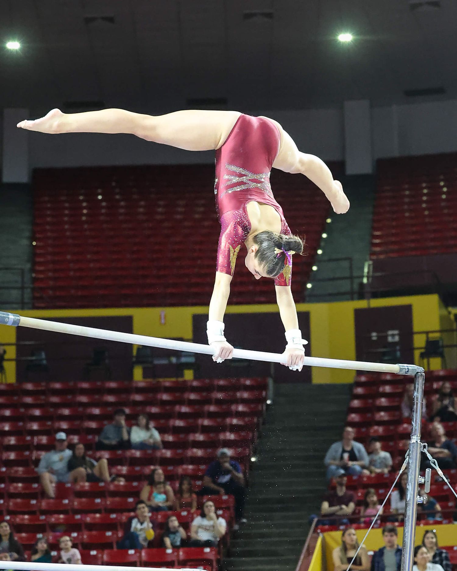 Female gymnast performing a release move on the uneven bars in a red leotard at an indoor competition.