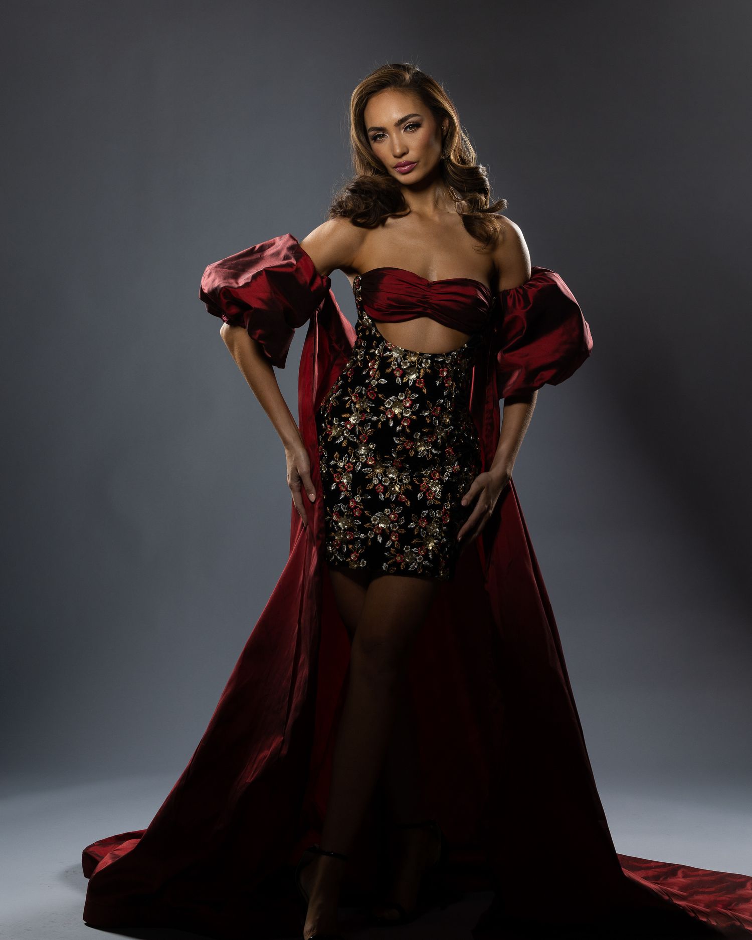 Woman in elegant red off-shoulder ballgown with floral embroidered skirt posing against gray studio background.