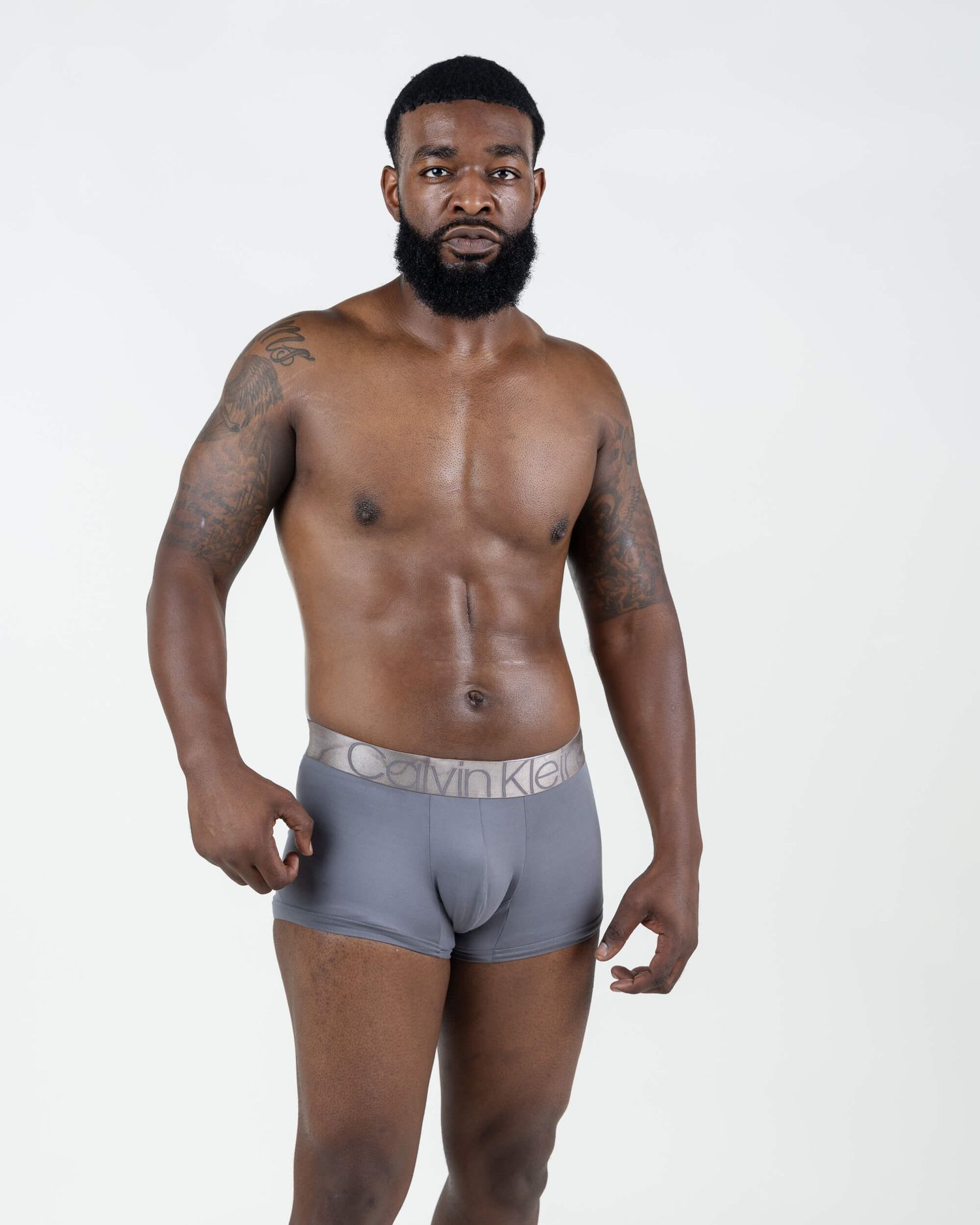 Man modeling gray Calvin Klein boxer briefs with tattoos on arms, standing against white background.
