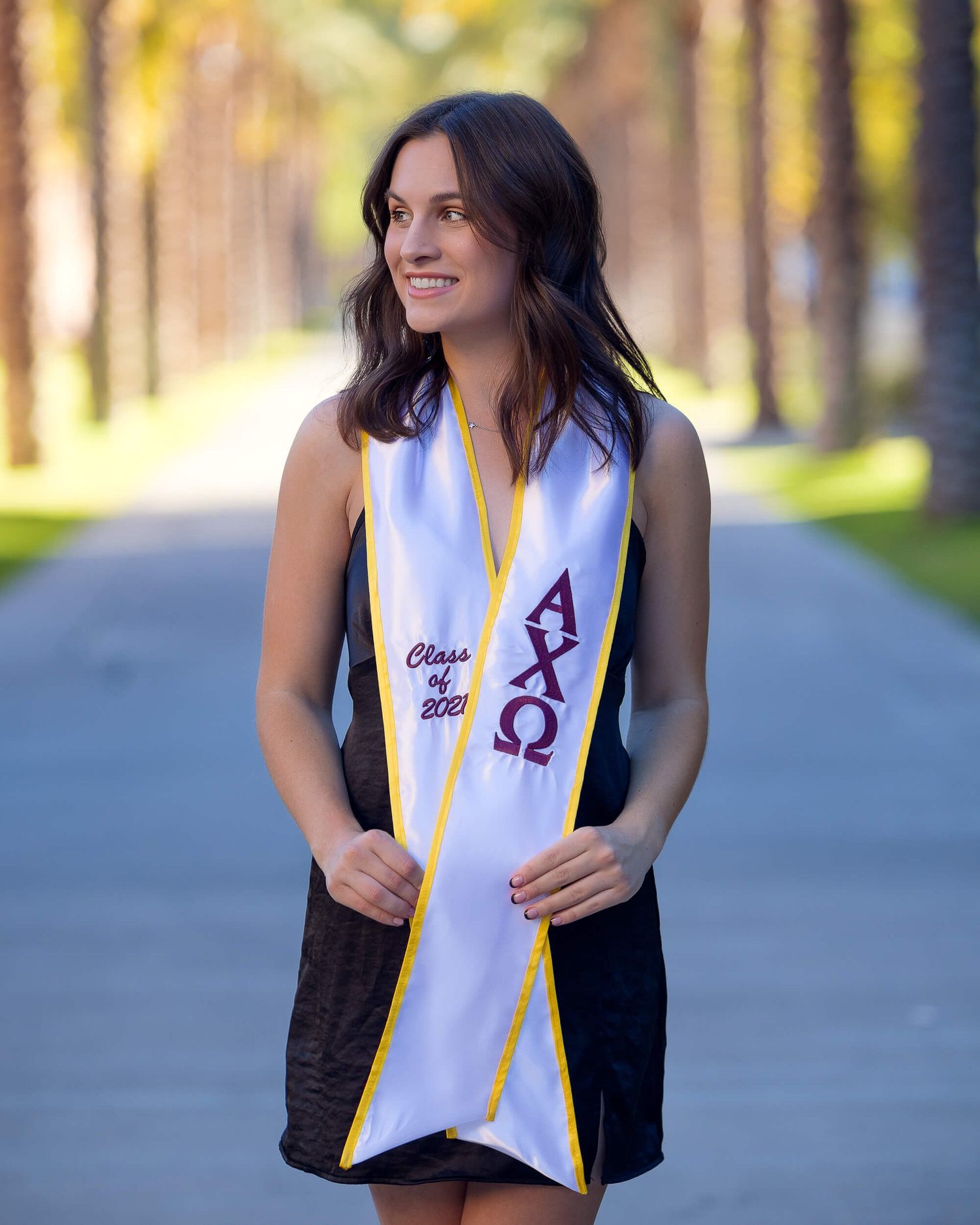 Young woman in black dress wearing Alpha Chi Omega Class of 2021 graduation stole on tree-lined campus path.