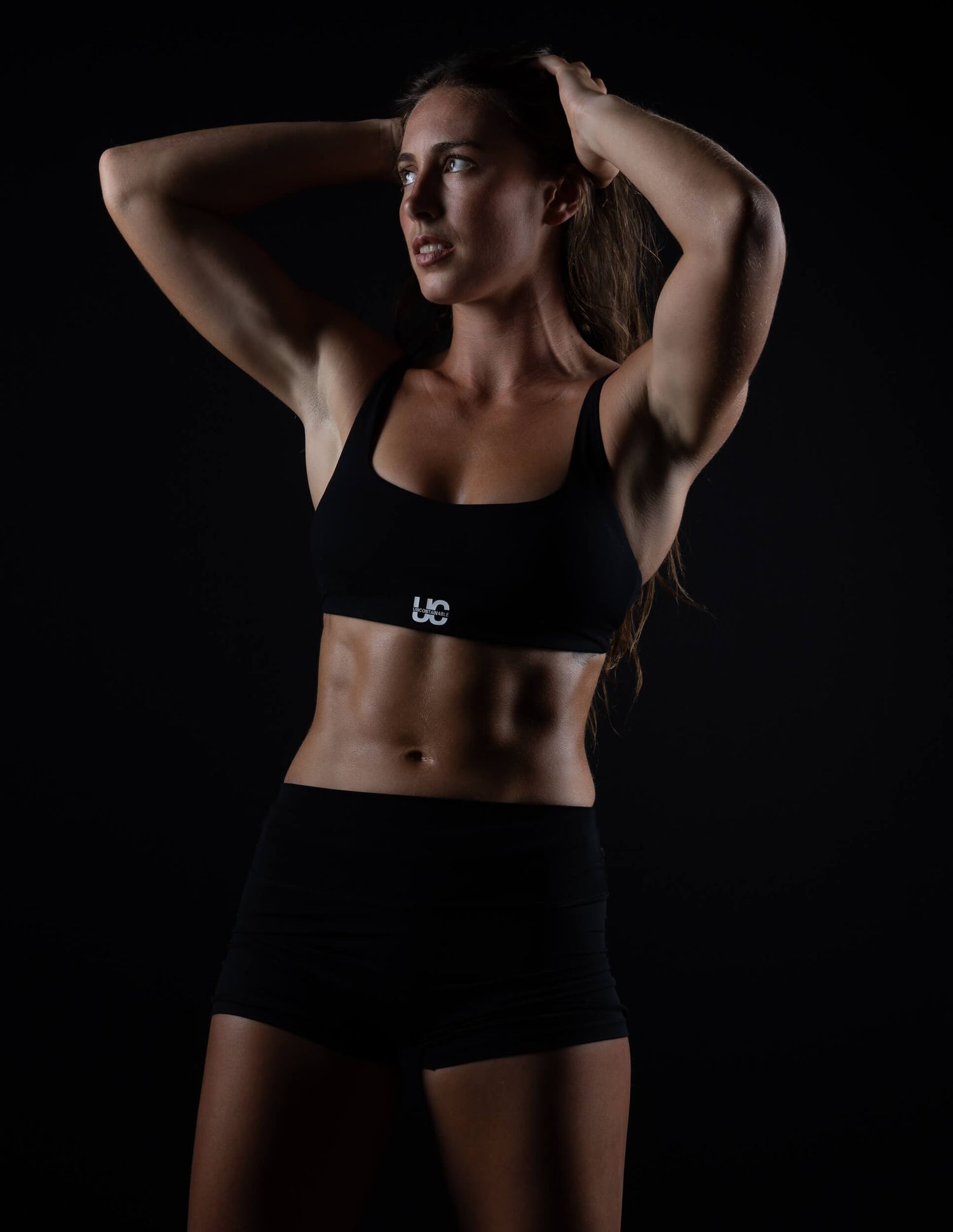 Fit woman in black sports bra and shorts posing with hands behind head in dark studio.