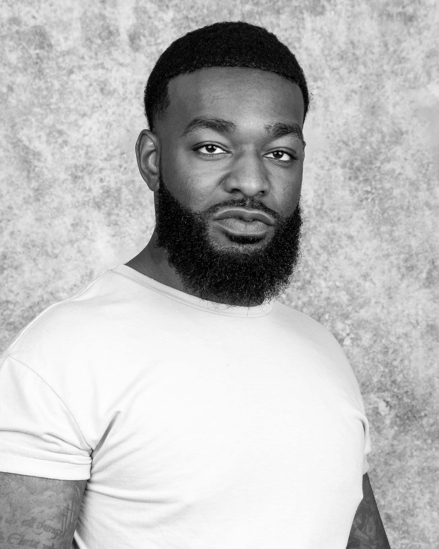 Black and white professional headshot of a bearded man in a white t-shirt against a textured backdrop.