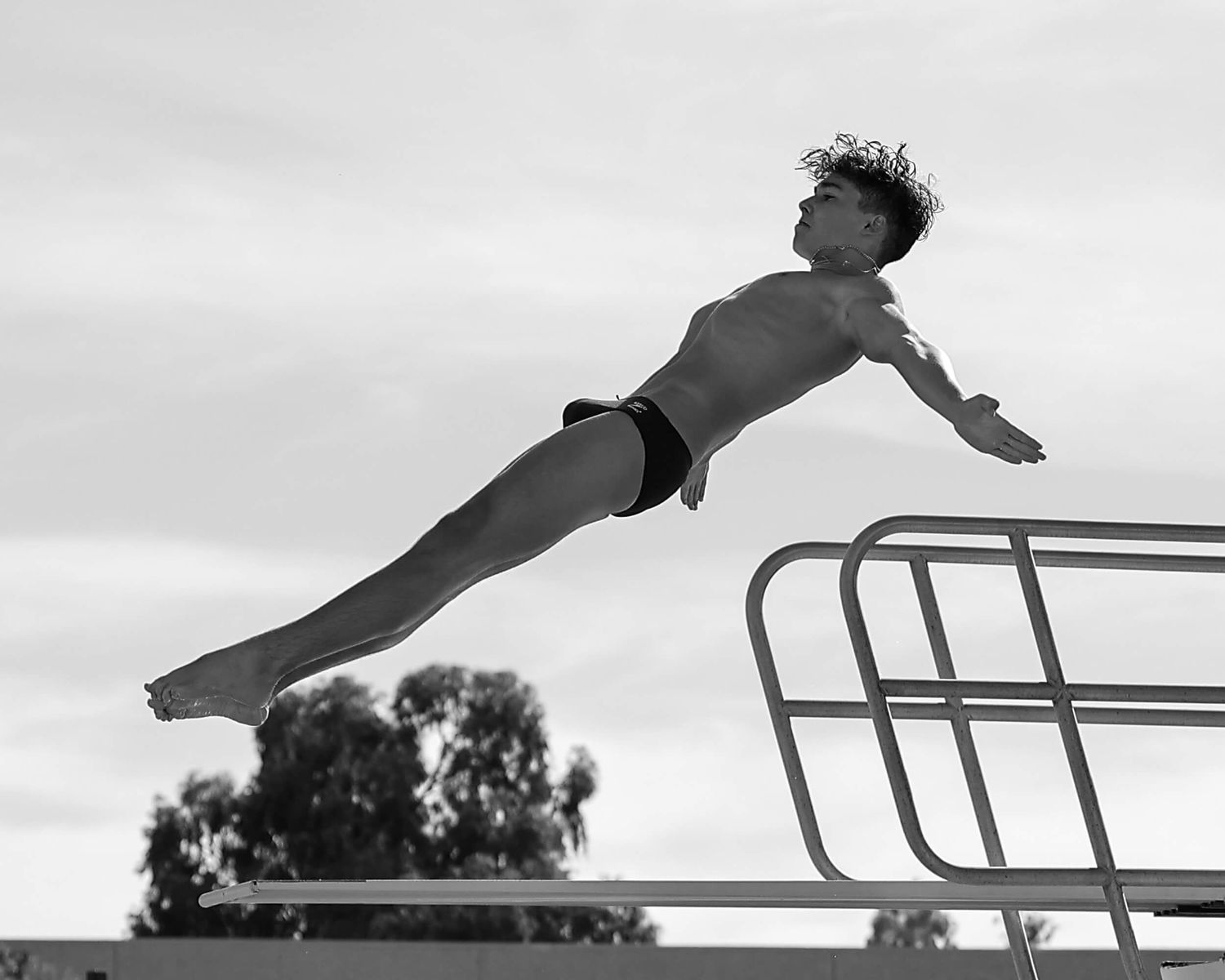 A male diver leaps gracefully from a diving board in a black and white action sports photo.