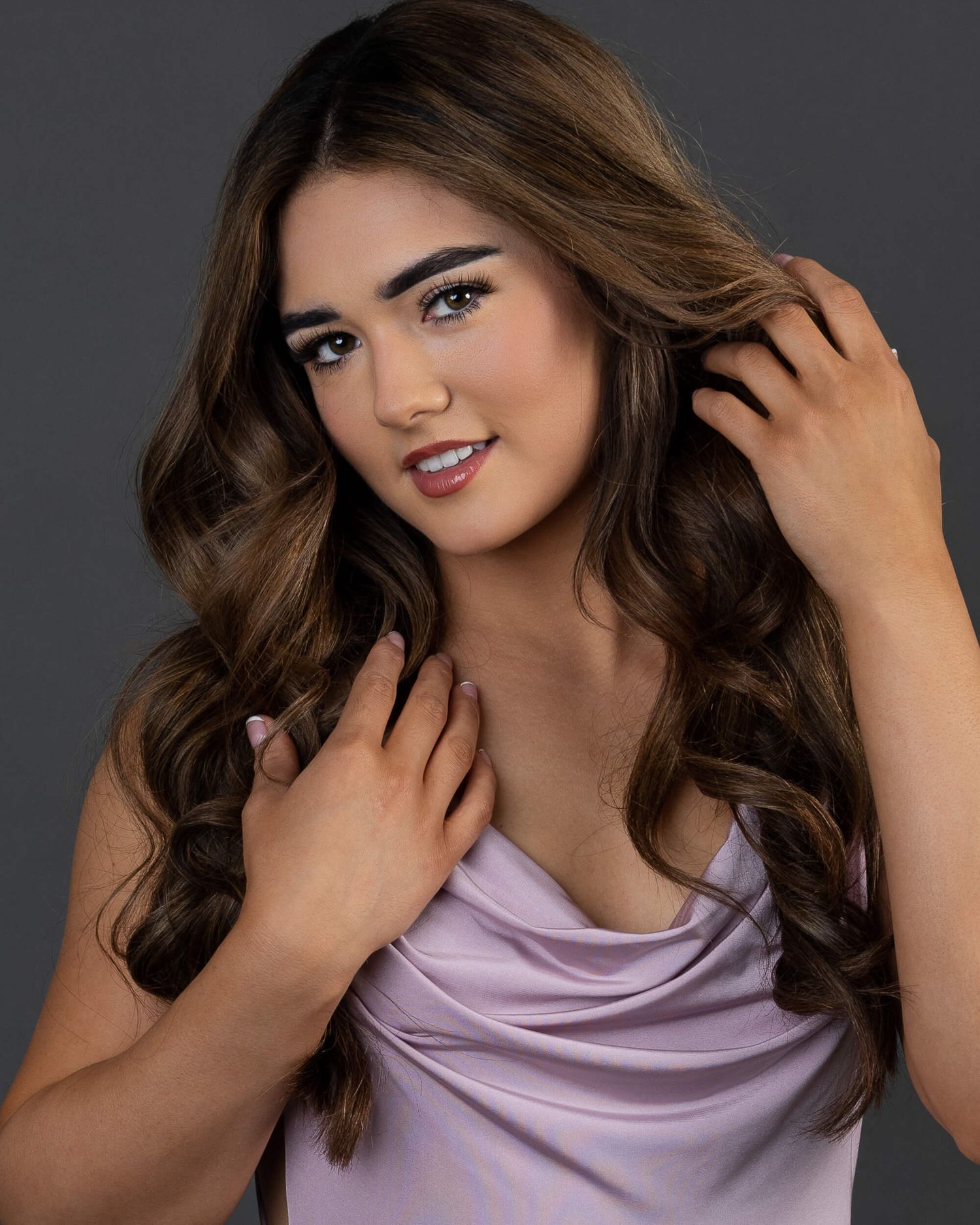 Young woman with long wavy brunette hair posing in a lavender satin top against a gray background.