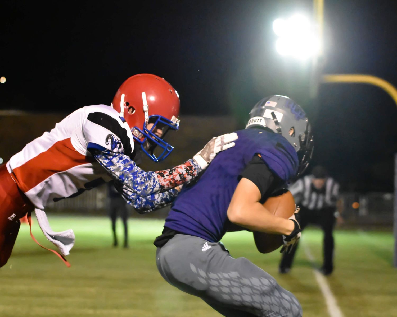 High school football player in red helmet tackles ball carrier under bright stadium lights during night game.