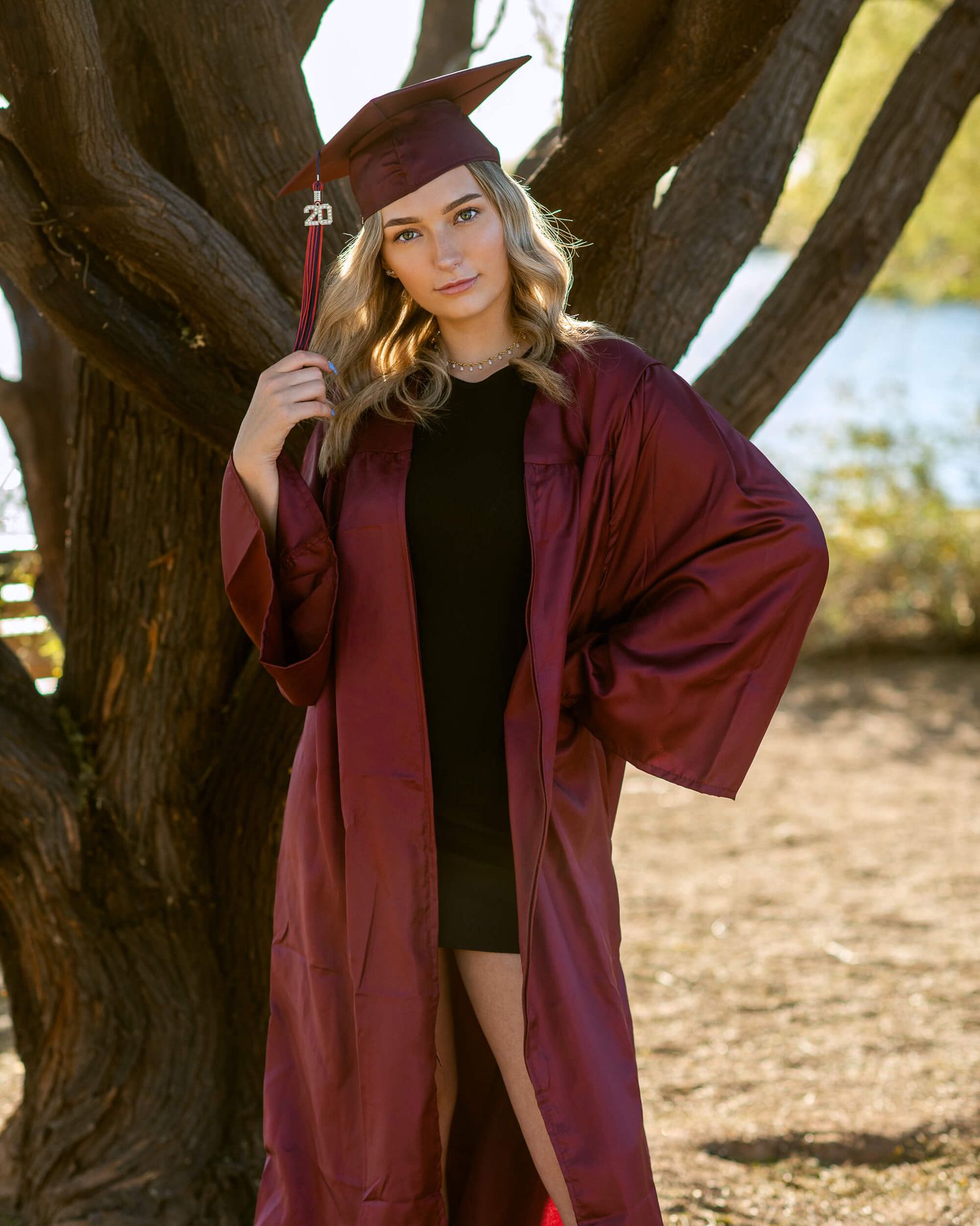Young woman in maroon graduation cap and gown poses confidently outdoors near a tree with a lake in the background.