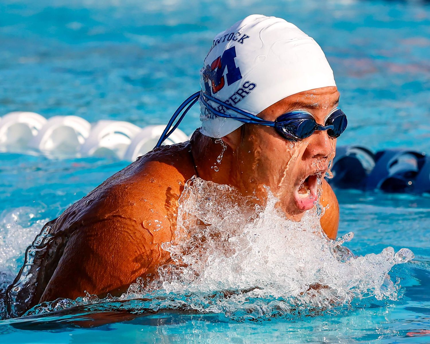 Competitive swimmer in white cap and goggles performs breaststroke in outdoor pool during race.