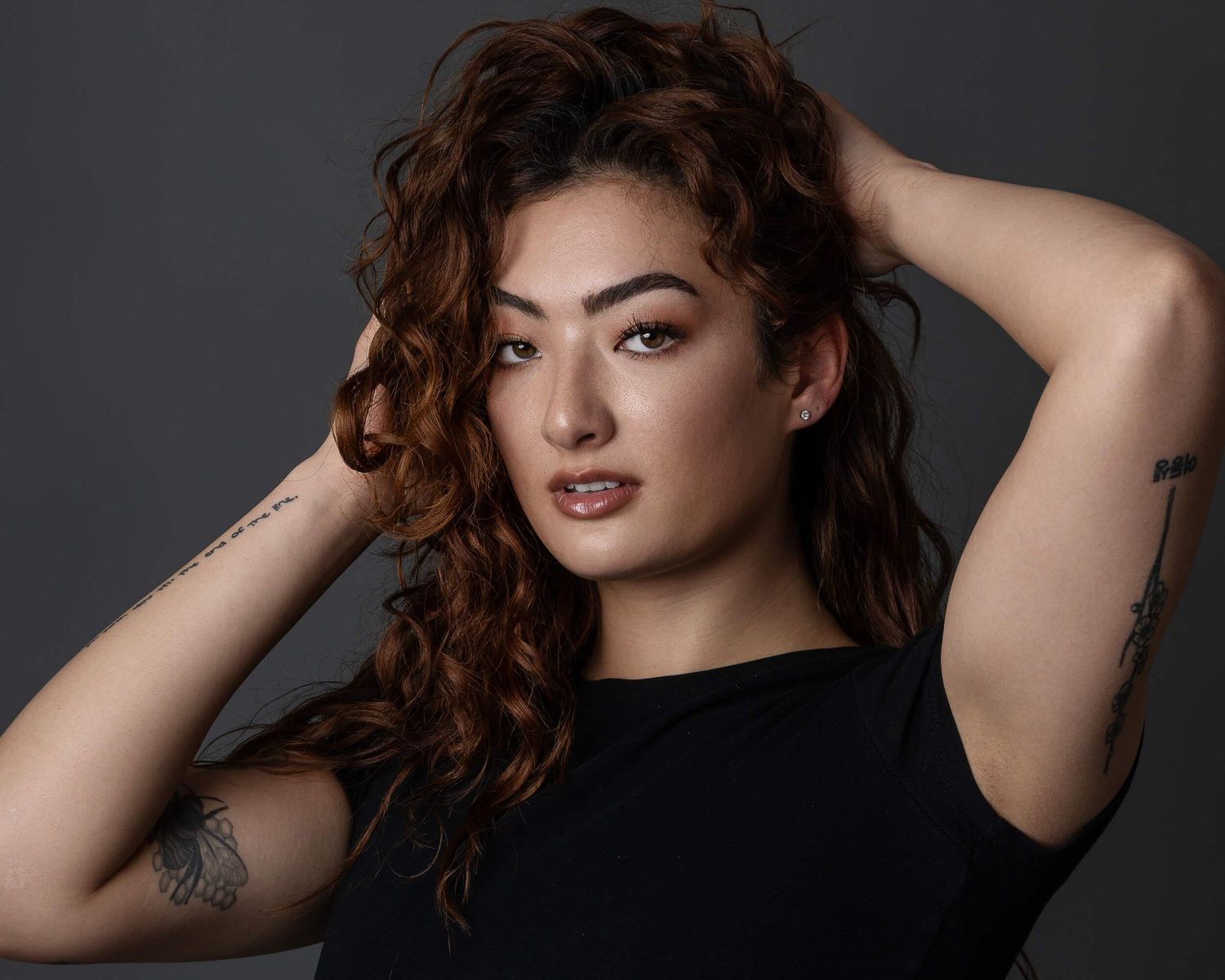 Young woman with curly auburn hair and tattoos posing in a black top against a dark background.