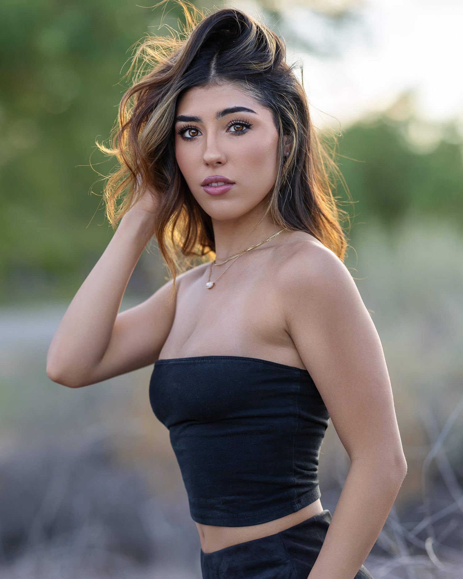 Young woman with highlighted hair in black tube top posing outdoors with bokeh background.