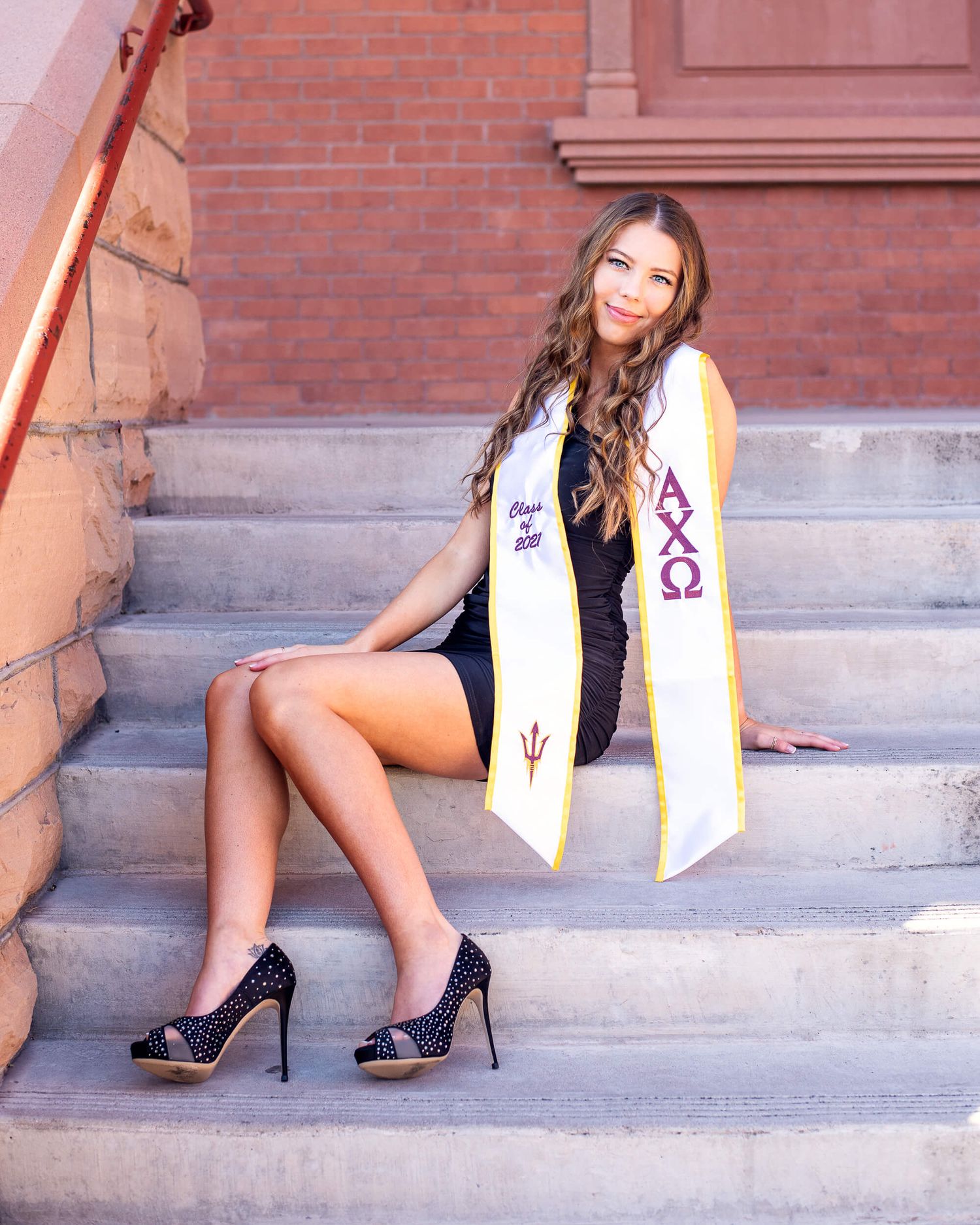 Young woman in black dress sitting on brick steps wearing Alpha Chi Omega graduation stole and heels.