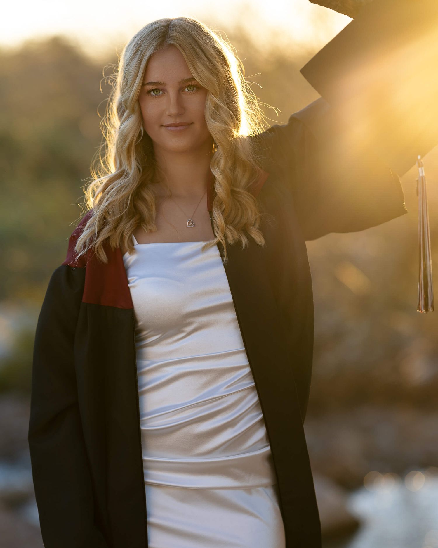 A smiling graduate in a black gown and white dress poses outdoors in golden sunset light holding a cap.