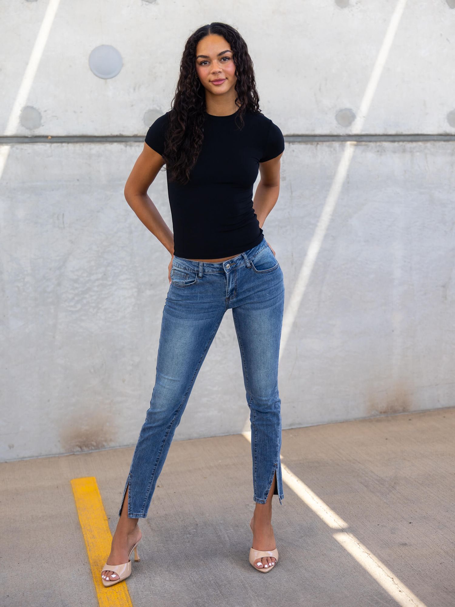 Woman in black tee and blue skinny jeans with split hem, wearing nude heels, posing confidently outdoors.