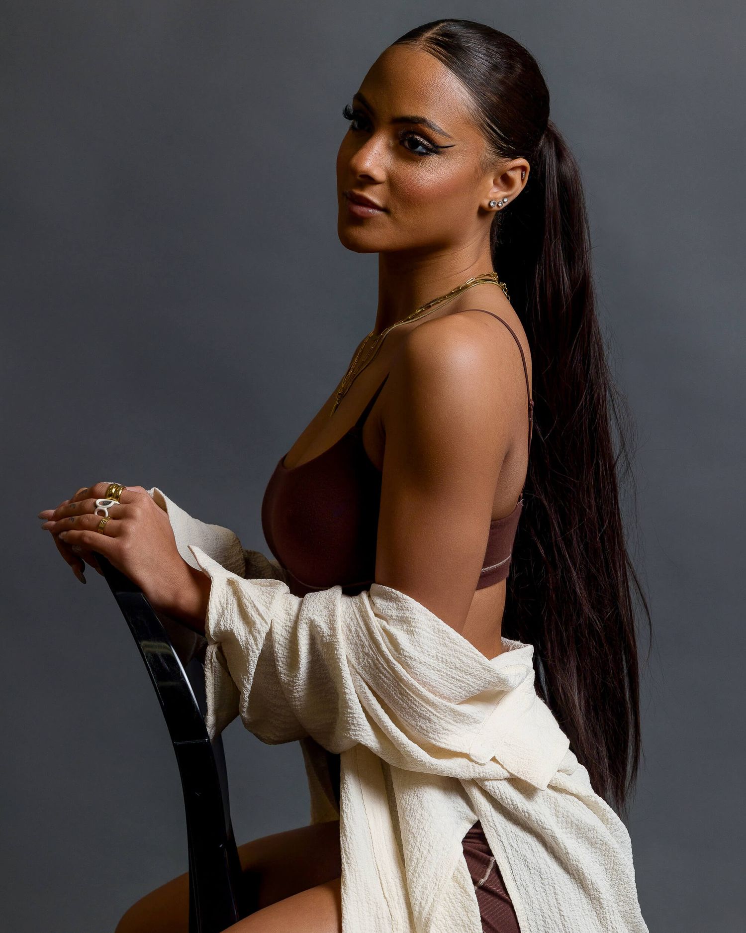 Woman with long sleek ponytail poses in brown bodysuit and cream cardigan on chair.