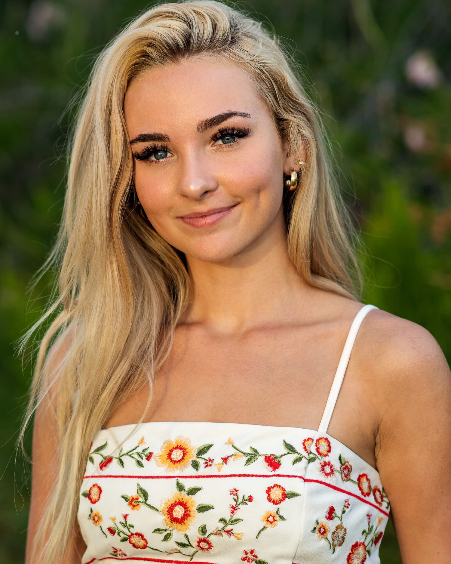 Young blonde woman smiling in a floral sundress with long straight hair in an outdoor setting.