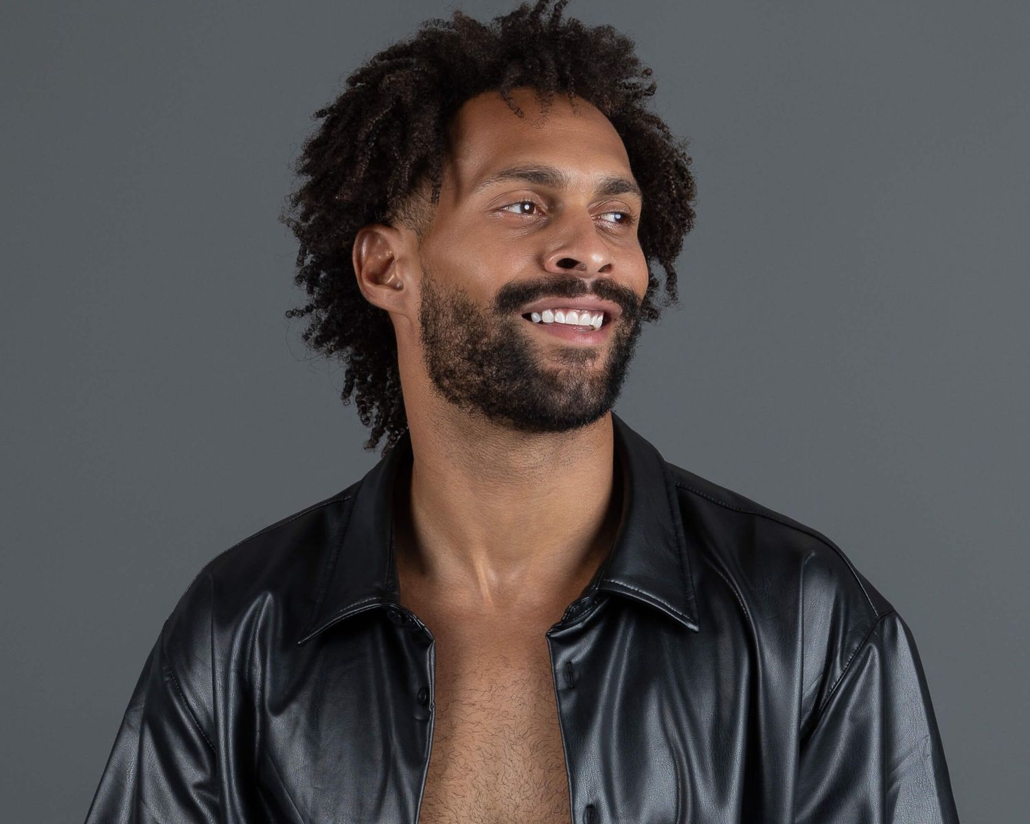 Smiling Black man with short curly hair wearing an open black leather jacket against a gray background.